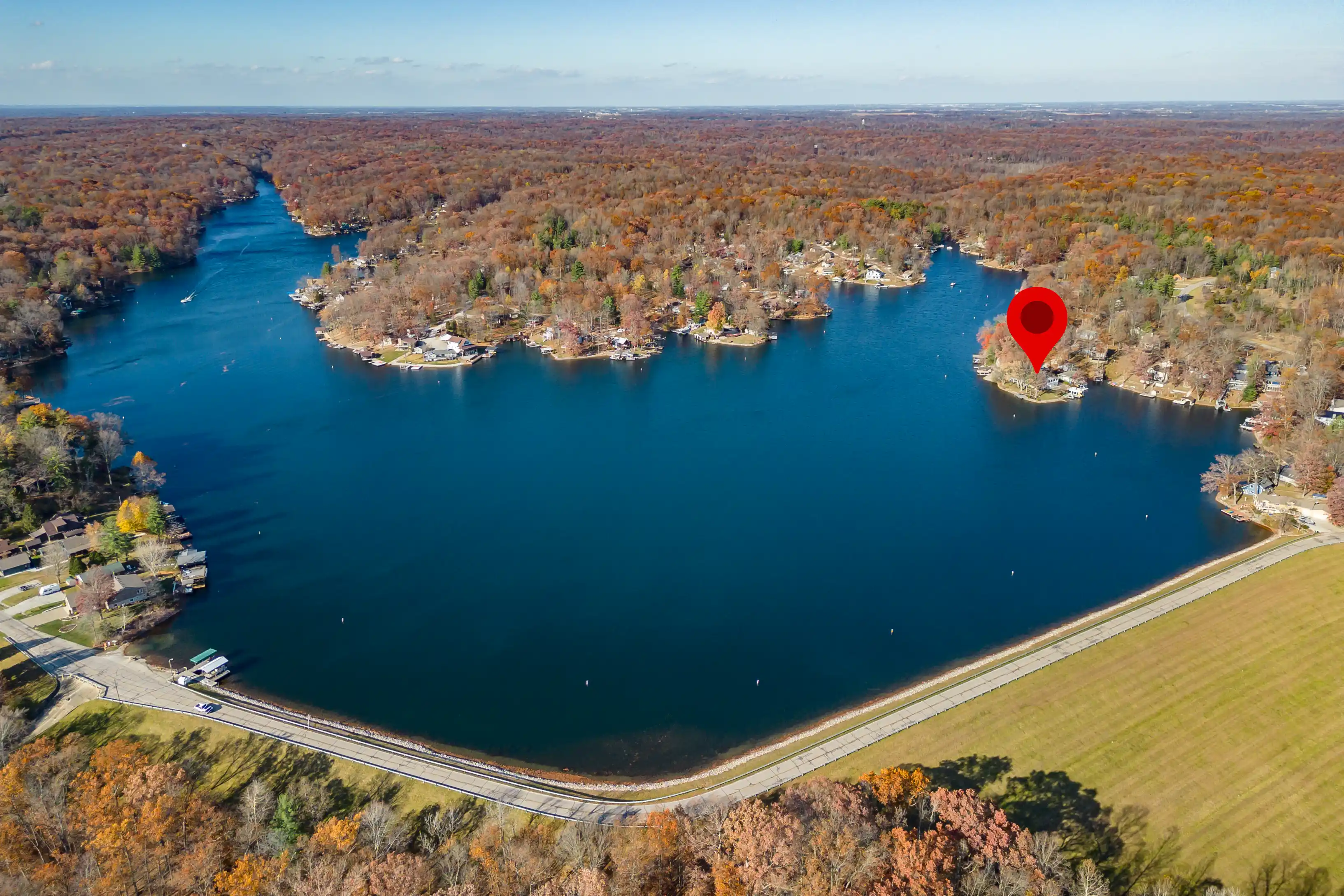Aerial view of a tranquil lake surrounded by autumn-colored trees with houses along the shoreline, a red location marker indicating a specific point on the peninsula.