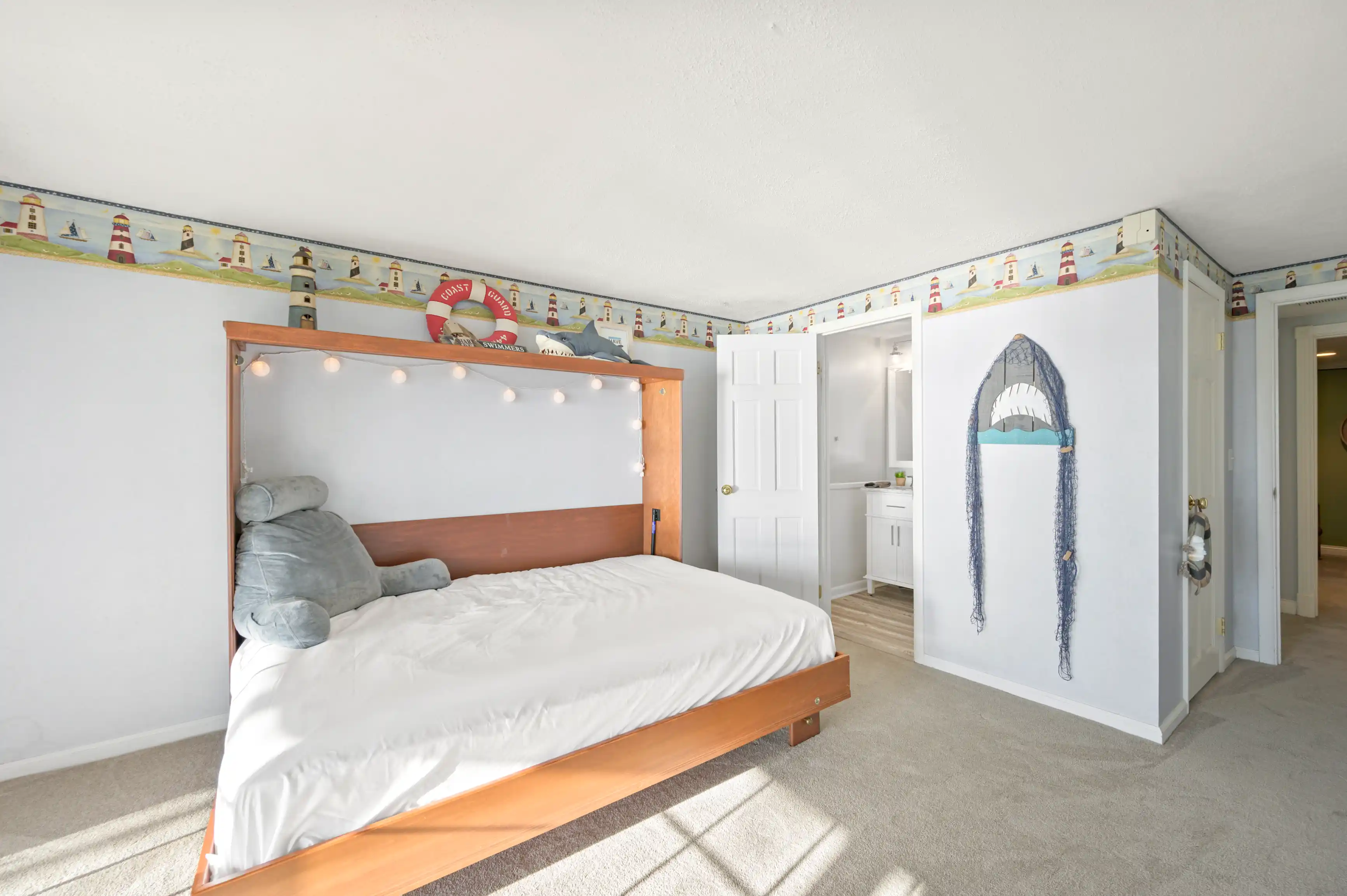Bright and airy bedroom with a nautical theme, featuring a bed with a wooden headboard adorned with string lights, plush manatee toy, lighthouse wallpaper border, and decorative shark silhouette on the closet door.