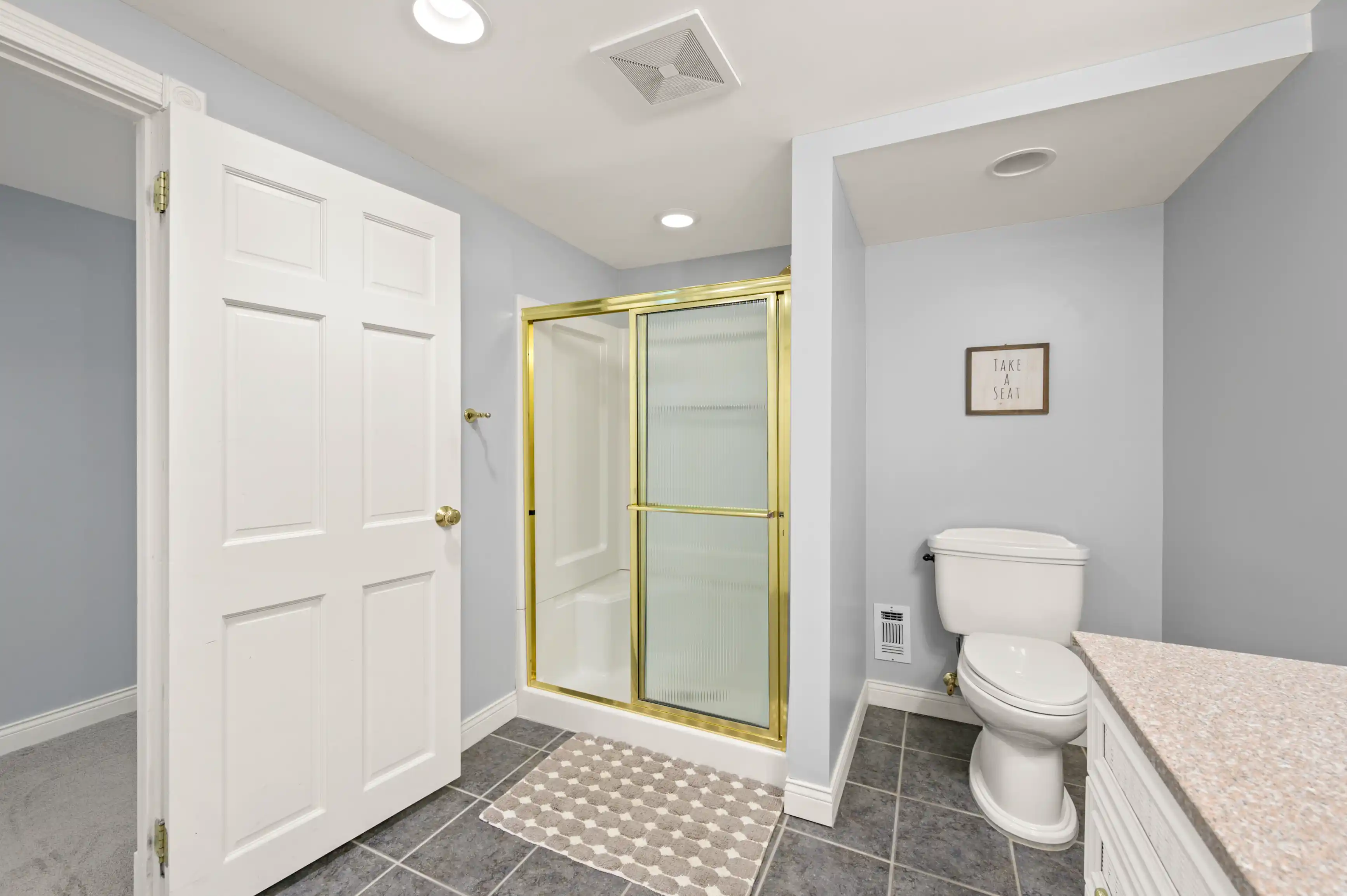 Modern bathroom interior with a closed white door, gray tiled floor, glass-enclosed shower with golden frame, white toilet, and a granite countertop against light blue walls with a decorative sign that reads "TAKE A SEAT".