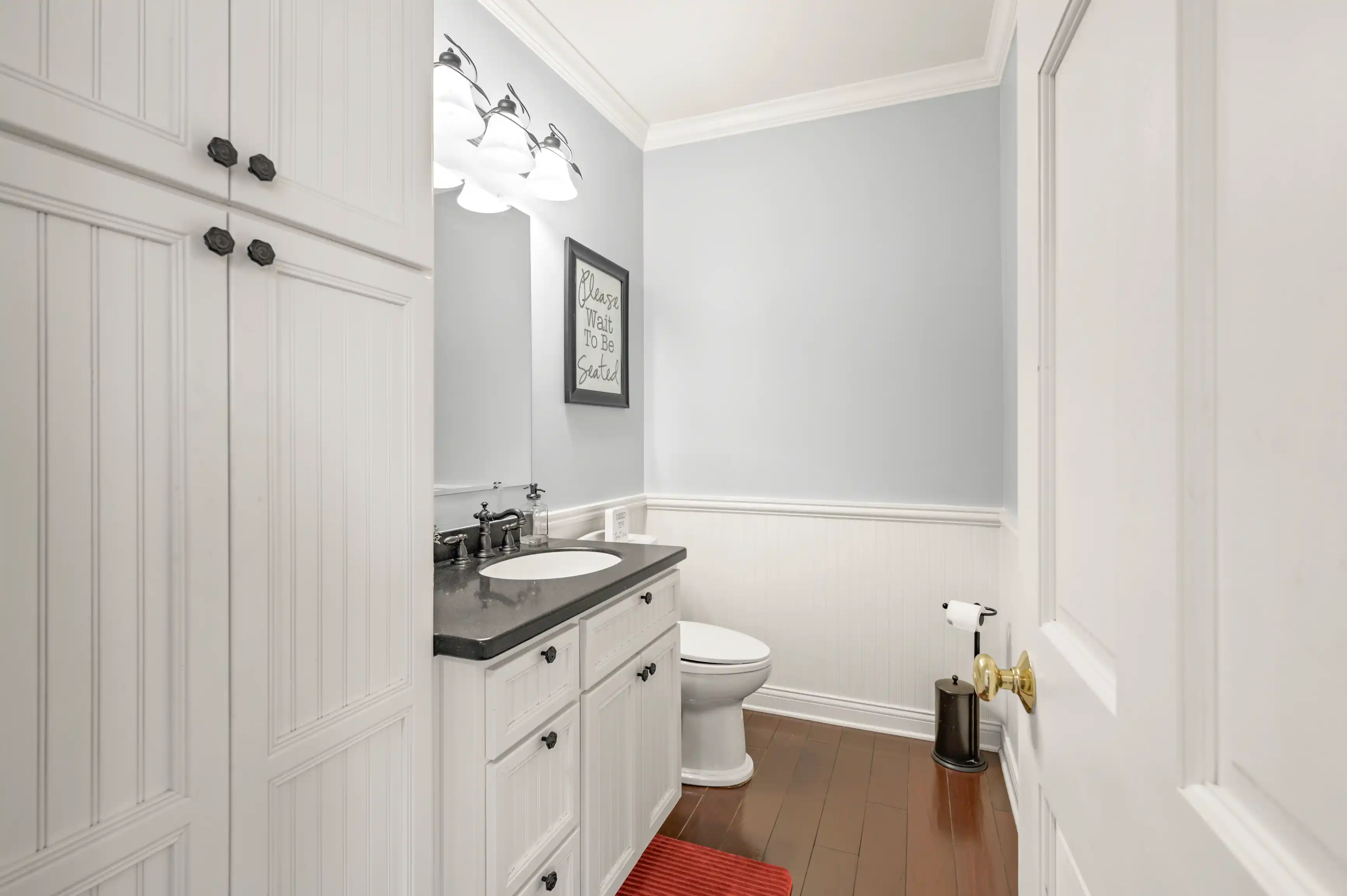 Interior of a clean bathroom with white cabinetry, a black countertop, an oval mirror, sconce lighting, and a decorative sign on the wall.
