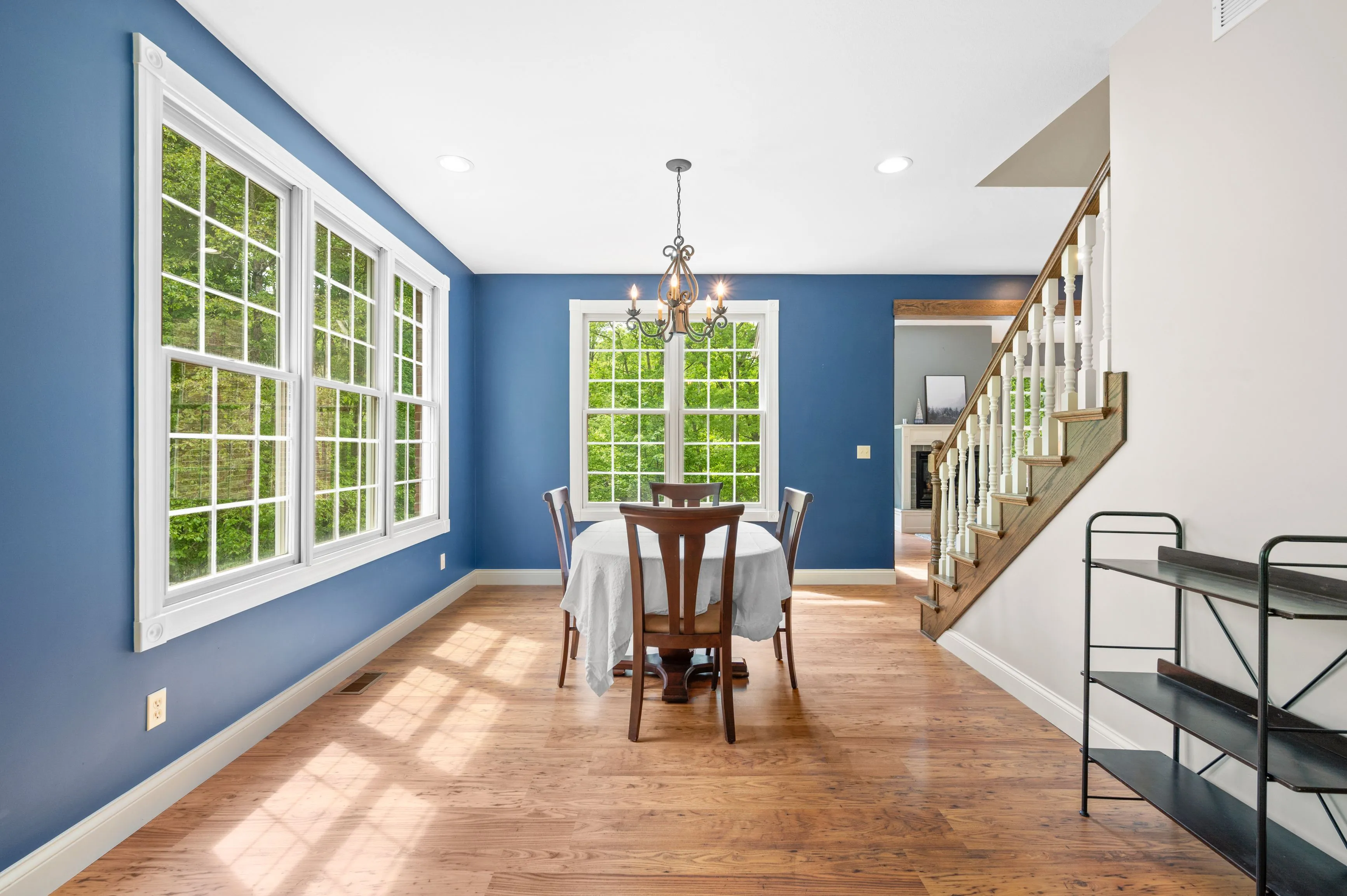 Bright dining room with blue walls, hardwood floors, large windows, a wooden dining table with chairs, and an adjacent staircase.
