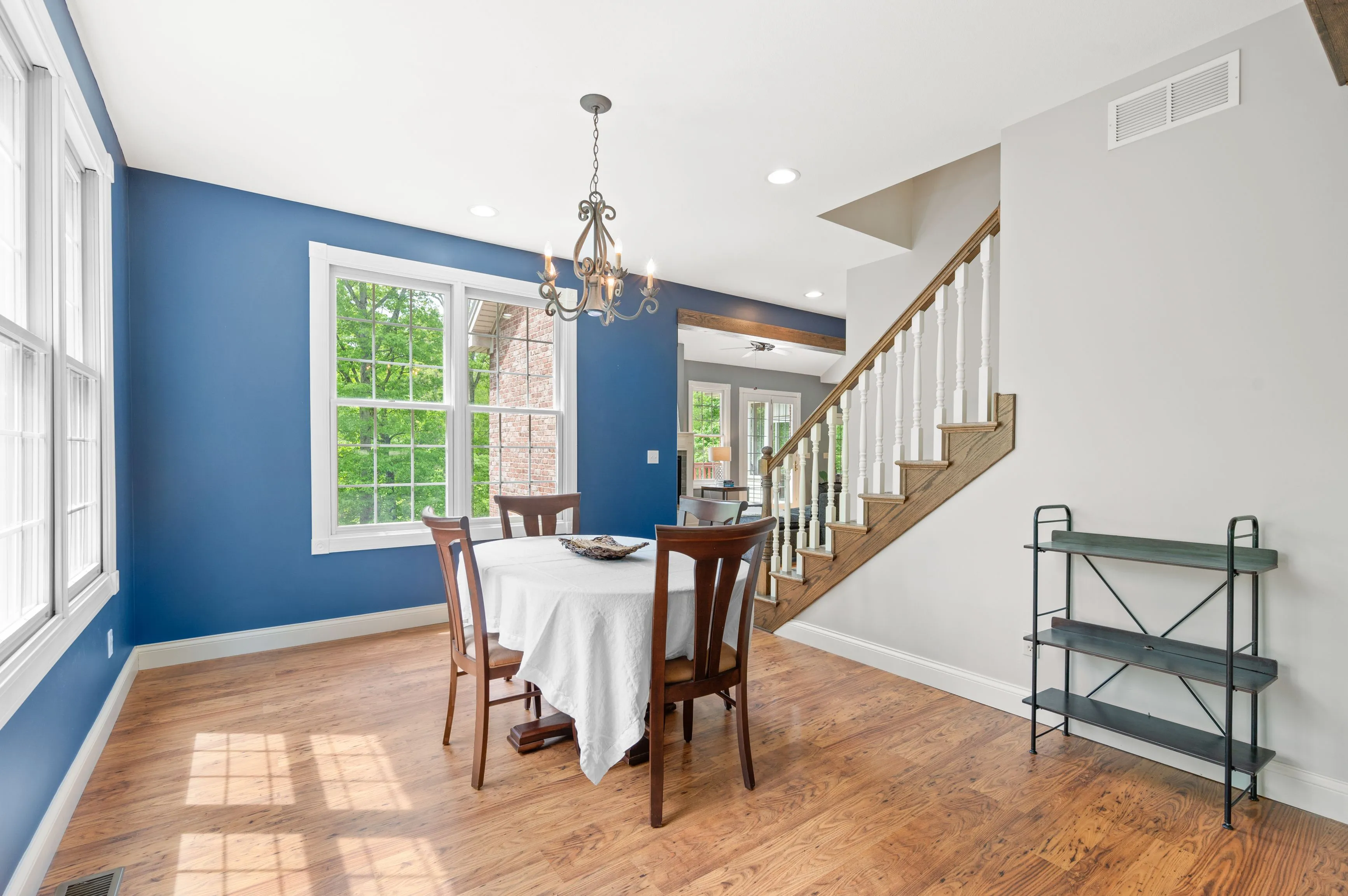 Bright dining room with blue walls, a wooden table set for four, hardwood floors, a classic chandelier, and a staircase leading to the upper level.