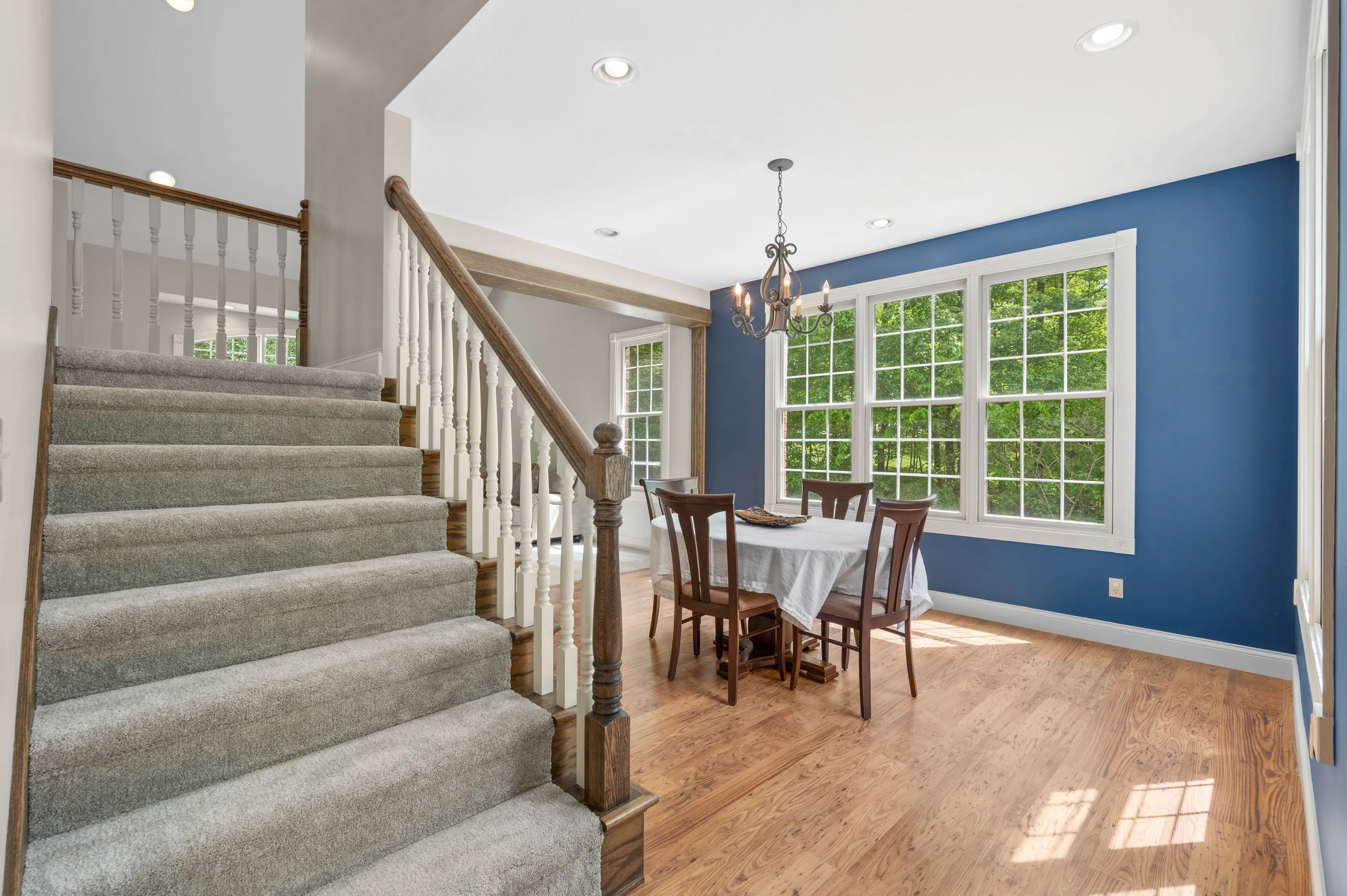 Elegant dining room with a blue accent wall, large windows, and a wooden table set adjacent to a staircase with white spindles and wooden banister.