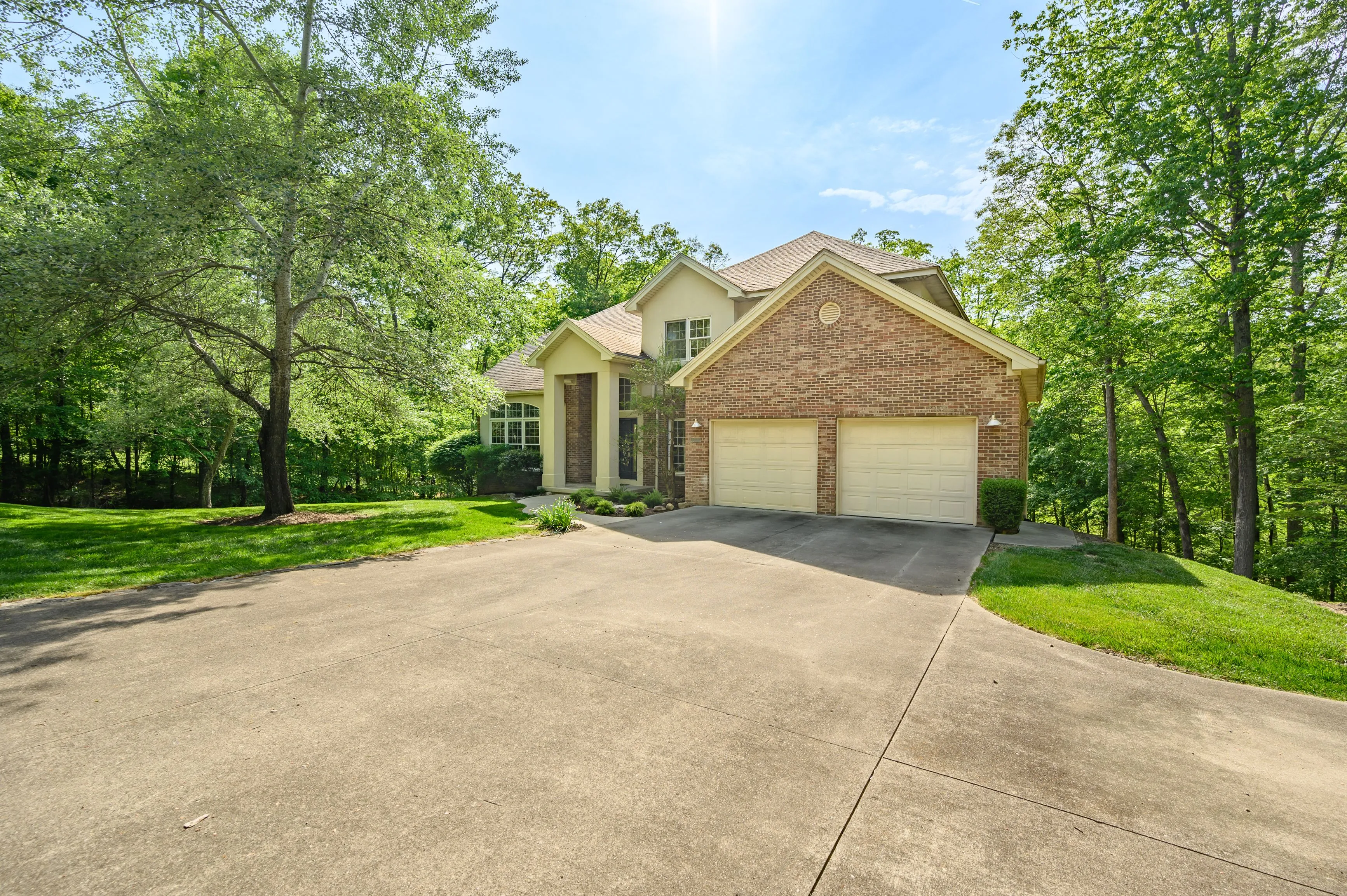 Brick house with double garage doors, surrounded by trees and a driveway, on a sunny day.