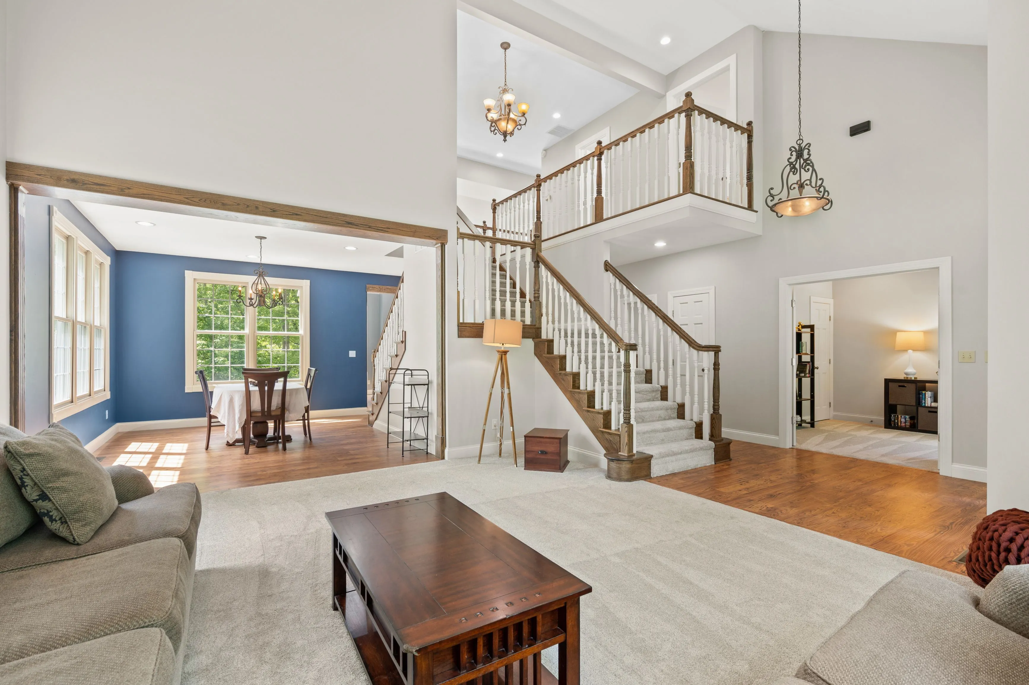 Spacious living room leading to an open dining area with a staircase to the second level, featuring hardwood floors and high ceilings.