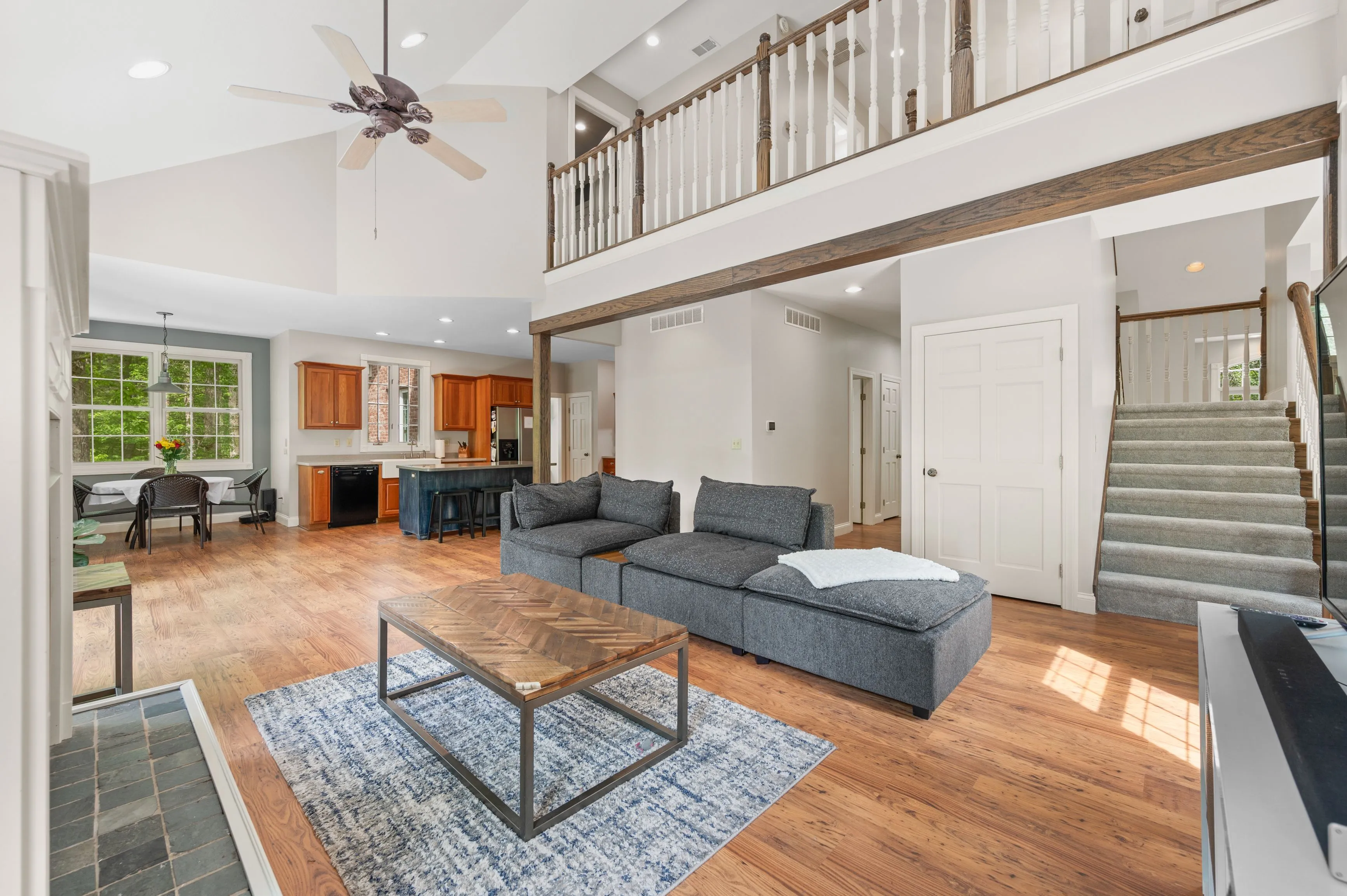 Bright and airy open-concept living space with a sectional sofa, hardwood floors, high ceiling with a fan, and an adjoining kitchen with wooden cabinets.