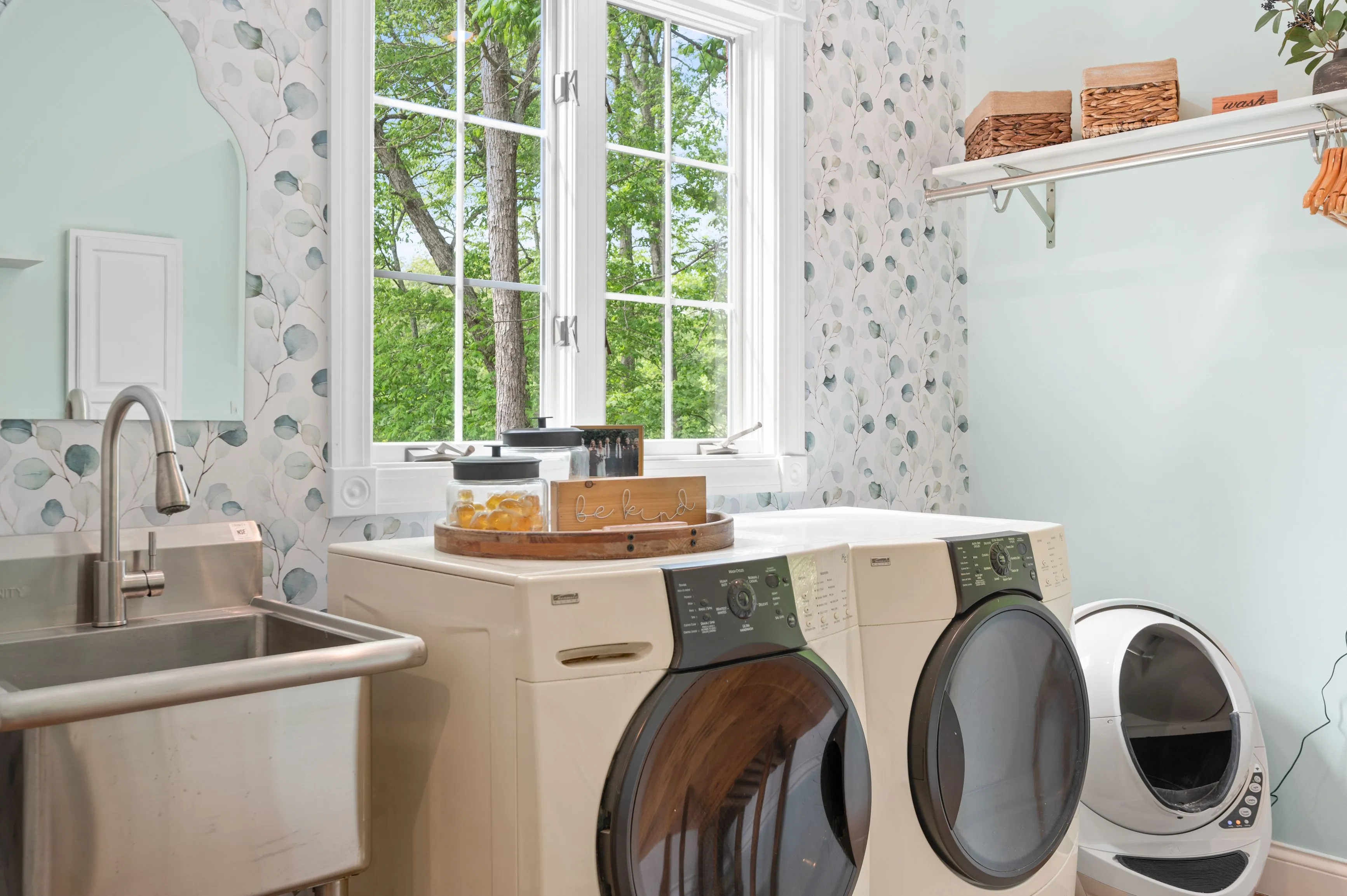 A bright laundry room with a front-loading washer and dryer, utility sink, and leaf-patterned wallpaper with a view of trees outside a window.