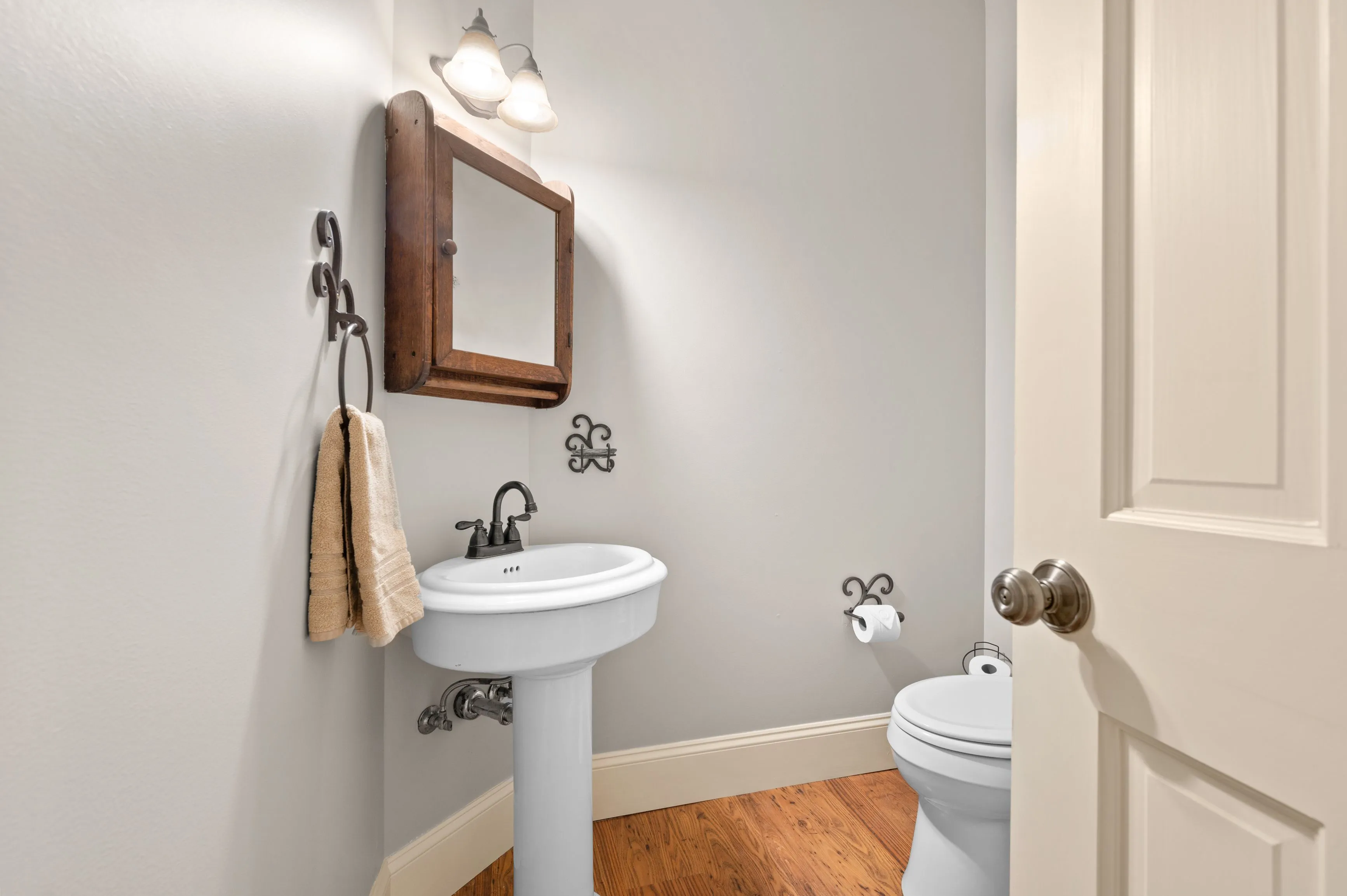 A small bathroom with wooden floors, white pedestal sink, toilet, beige towel on a hook, wooden mirror, and wall-mounted lights.