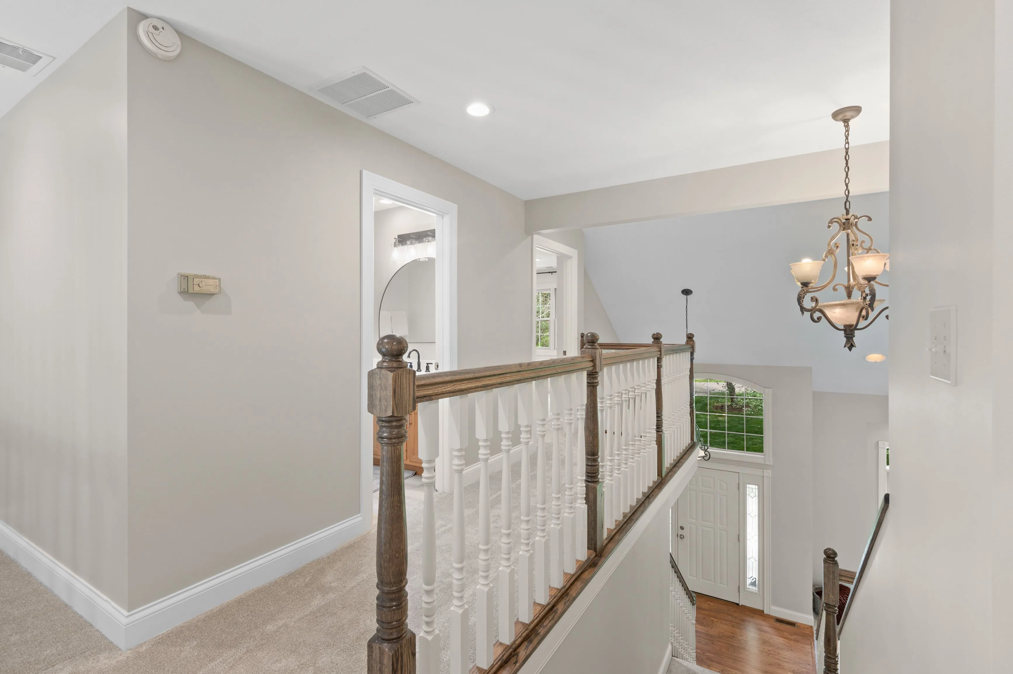 Bright upper landing area with beige walls, a wooden balustrade, and a classic chandelier overlooking an entrance with a white door.
