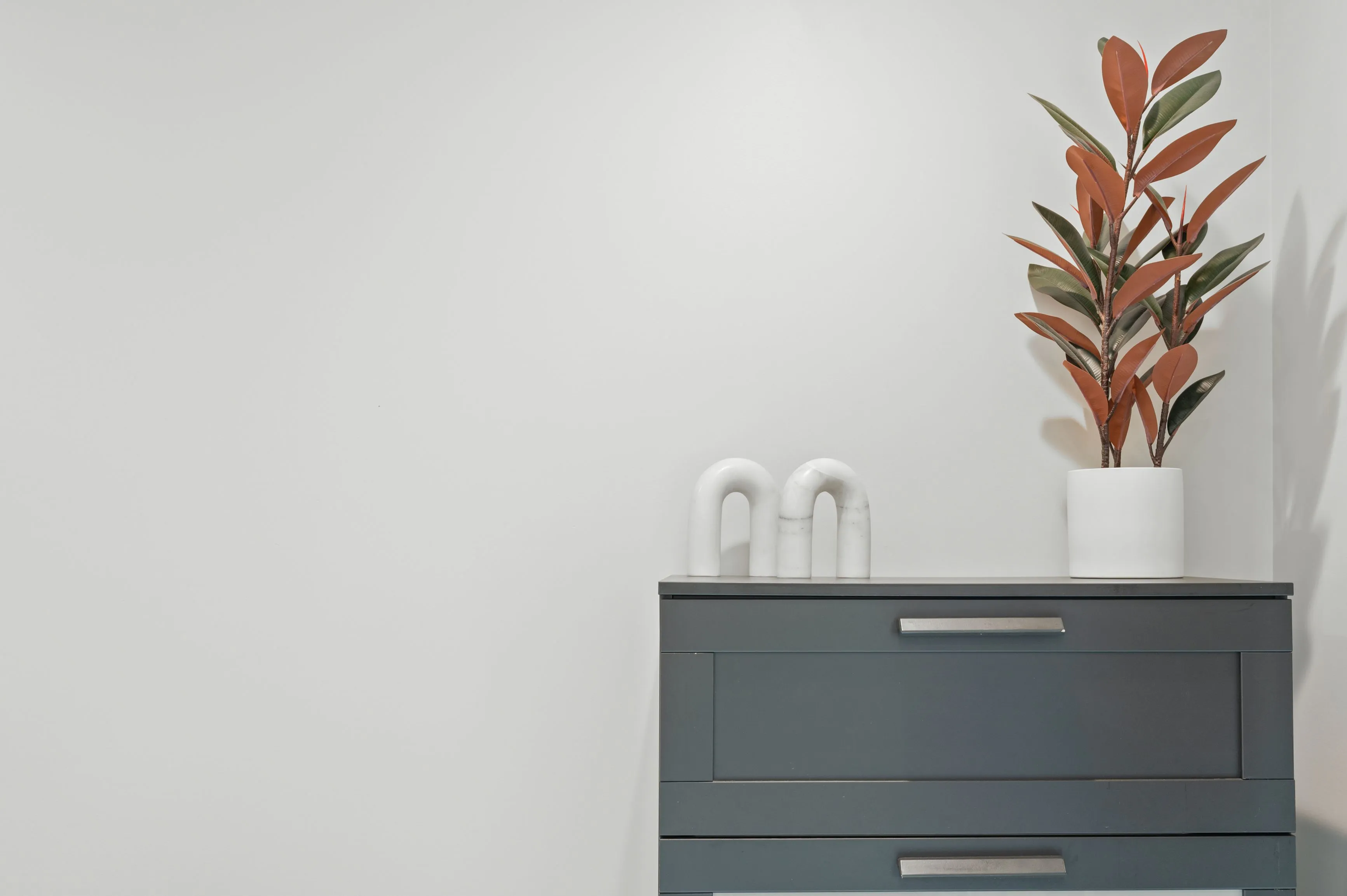 A minimalist room with a dark grey chest of drawers on the right, topped with a white vase holding a red and green leafy plant, and a pair of white decorative arches.