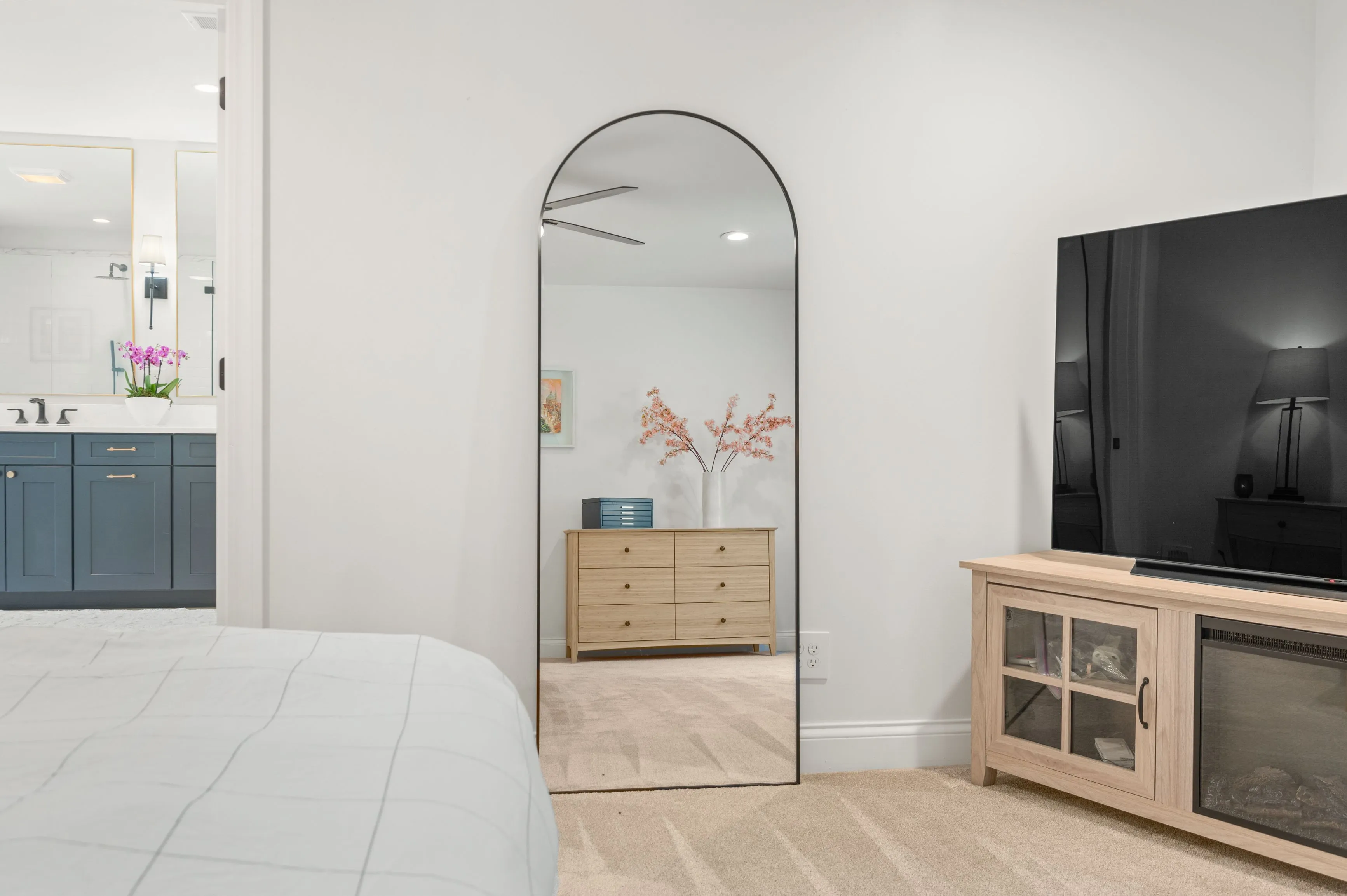 Modern bedroom interior with a wooden dresser, arch mirror, television and a glimpse into the adjoining bathroom with blue cabinets and pink flowers on the vanity.