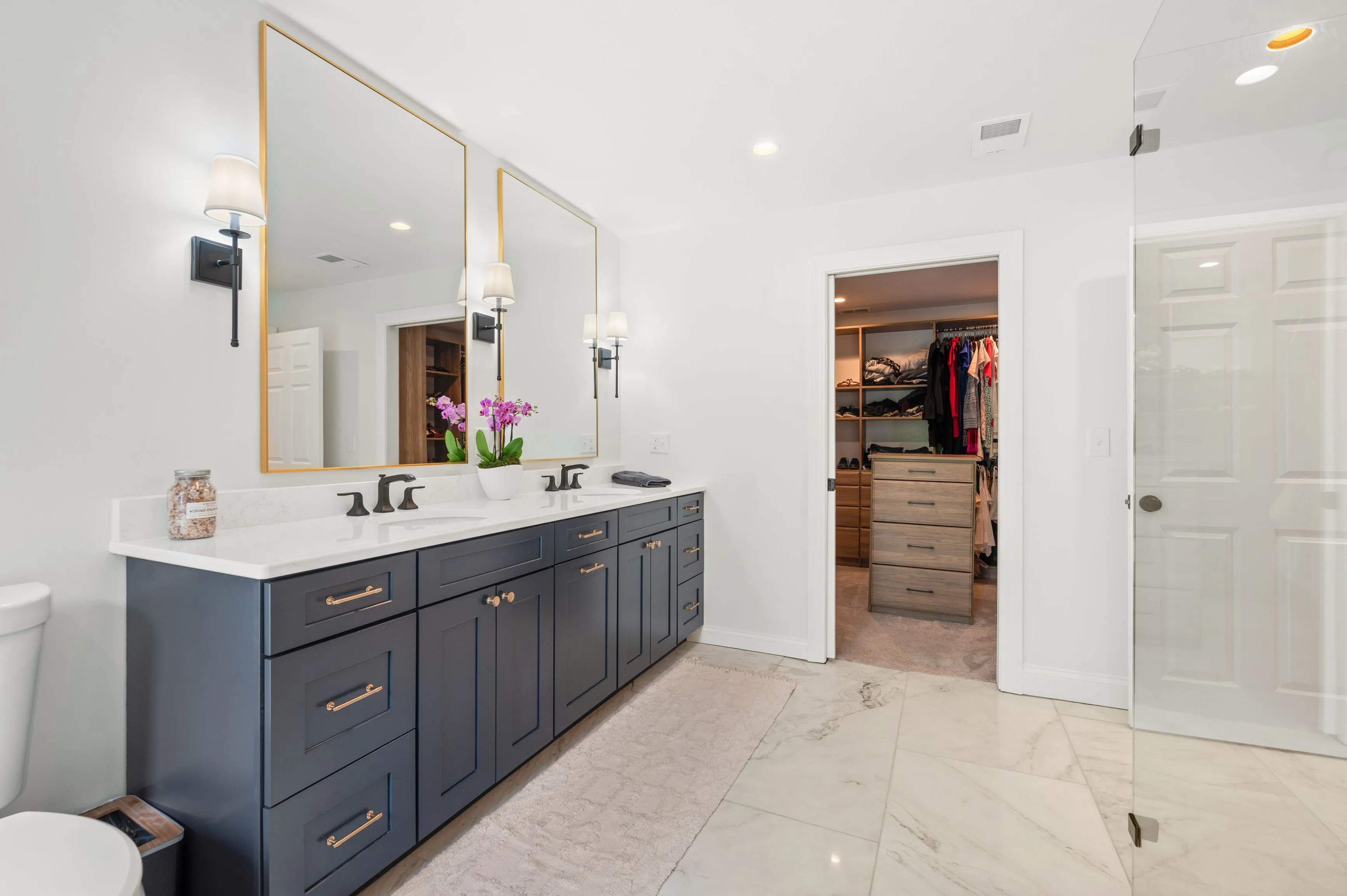 Modern bathroom interior with a double vanity cabinet in navy blue, white marble countertop, large mirror with gold frame, and a walk-in closet visible in the background.