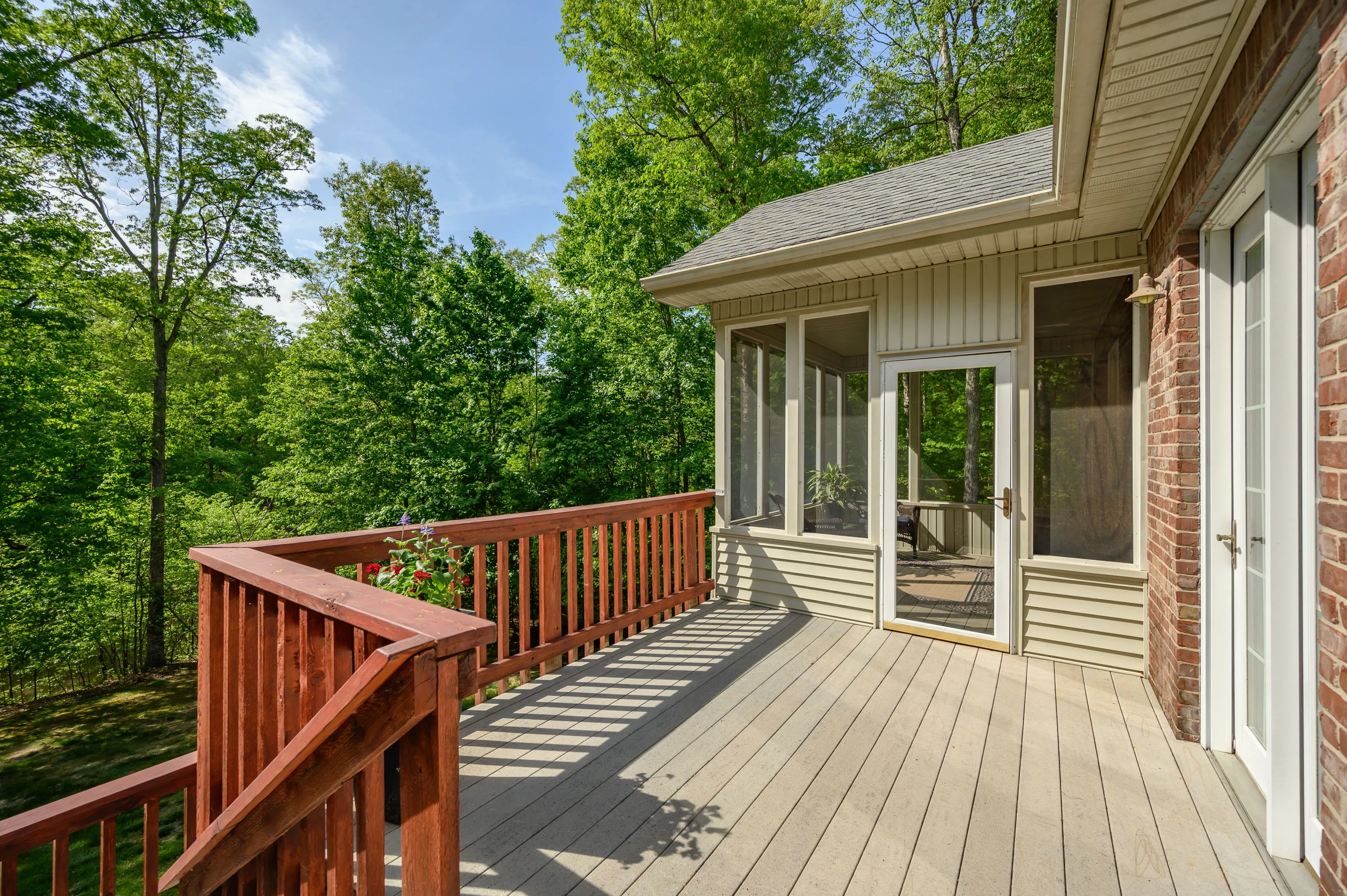Wooden deck of a house with railing overlooking a lush green forest with a screened porch area to the side.