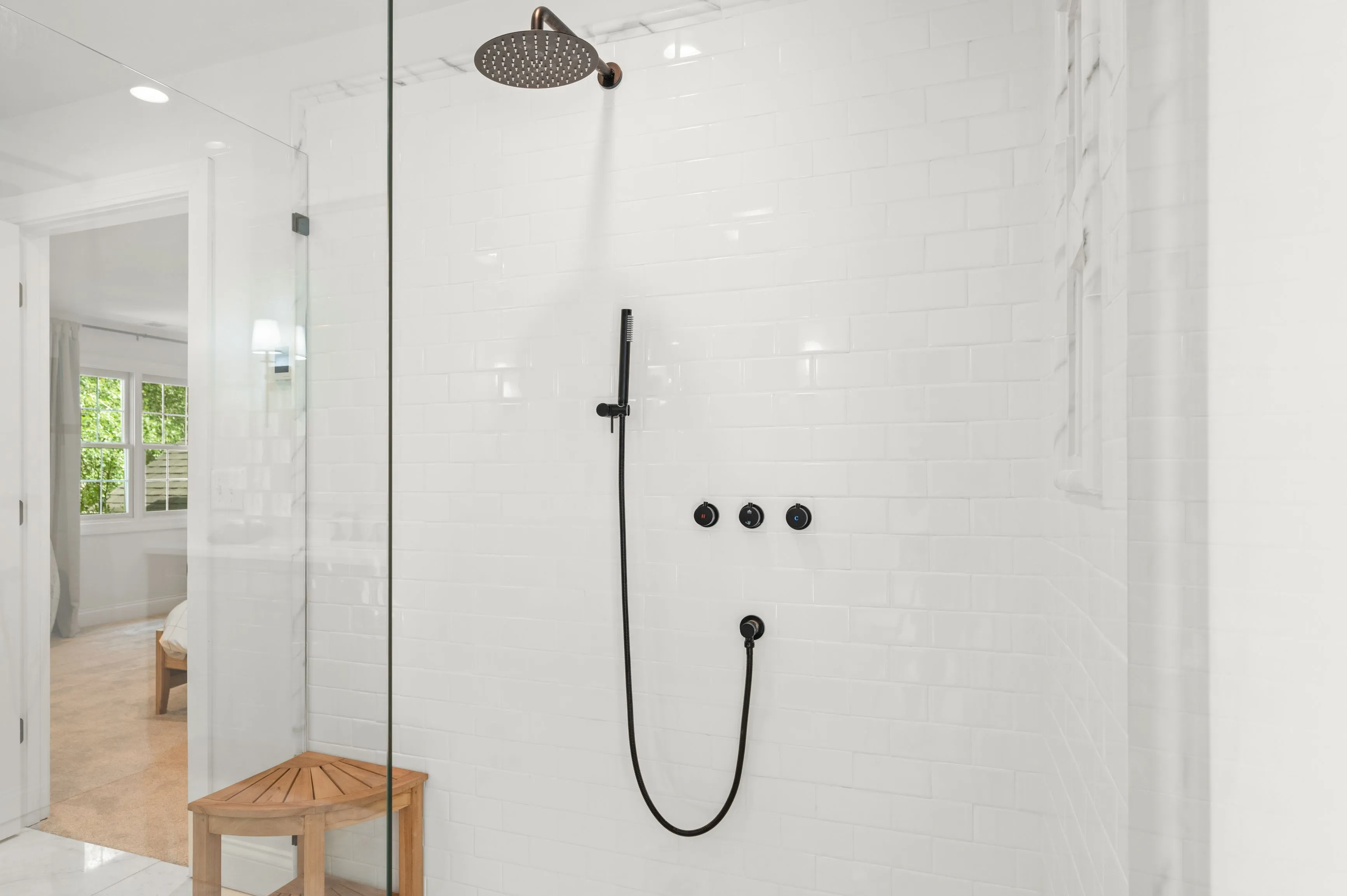 Modern shower with white subway tiles, glass door, bronze shower head and fixtures, and a wooden stool.