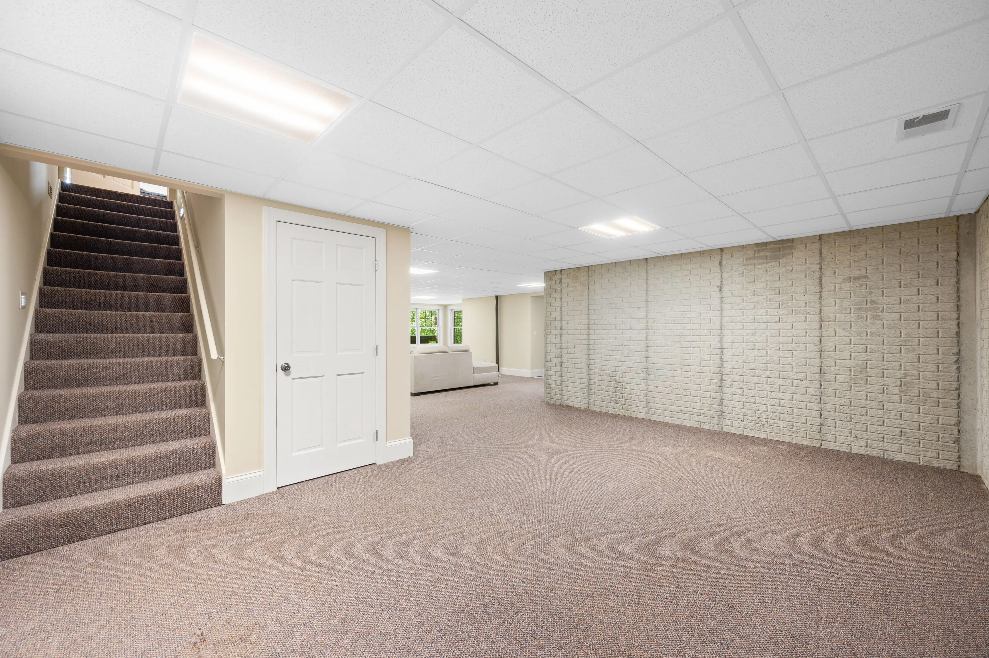 A spacious basement room with beige walls, carpeted floor, a white door, a set of stairs, and recessed lighting.