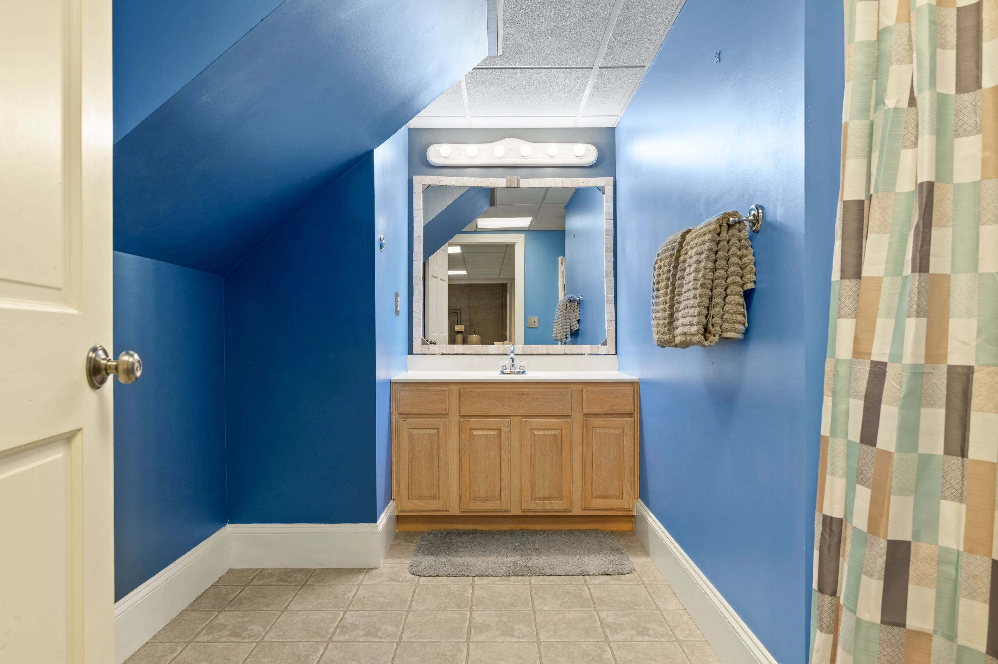 A bright blue bathroom featuring a wooden vanity with a large mirror, tiled flooring, and a plaid shower curtain.