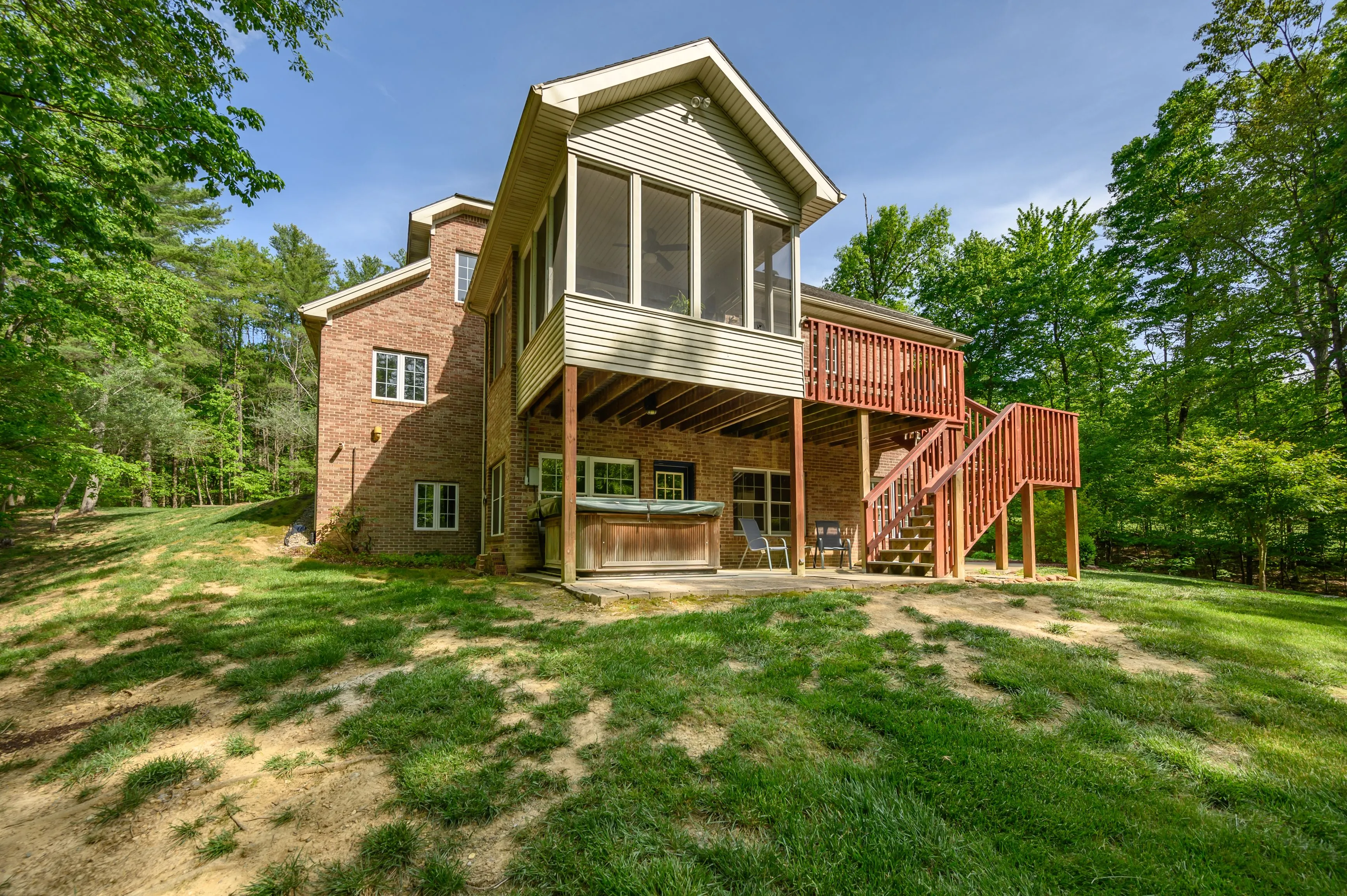 Brick house with screened upper deck, red wooden stairs, and hot tub, surrounded by trees and lawn on a sunny day.