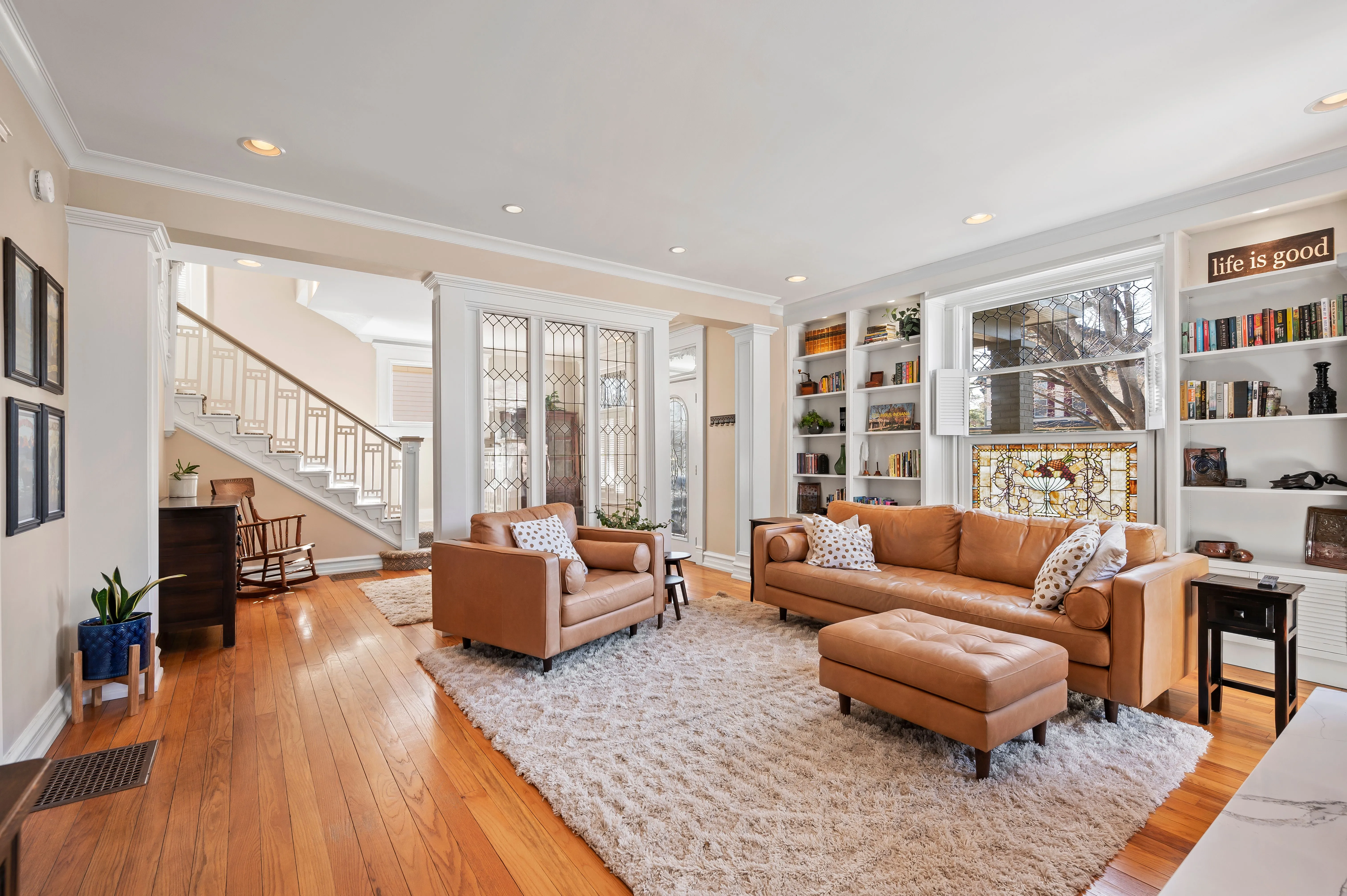 Bright and spacious living room interior with tan sofas, a large area rug, hardwood floors, built-in bookshelves, and staircase leading to the upper level.
