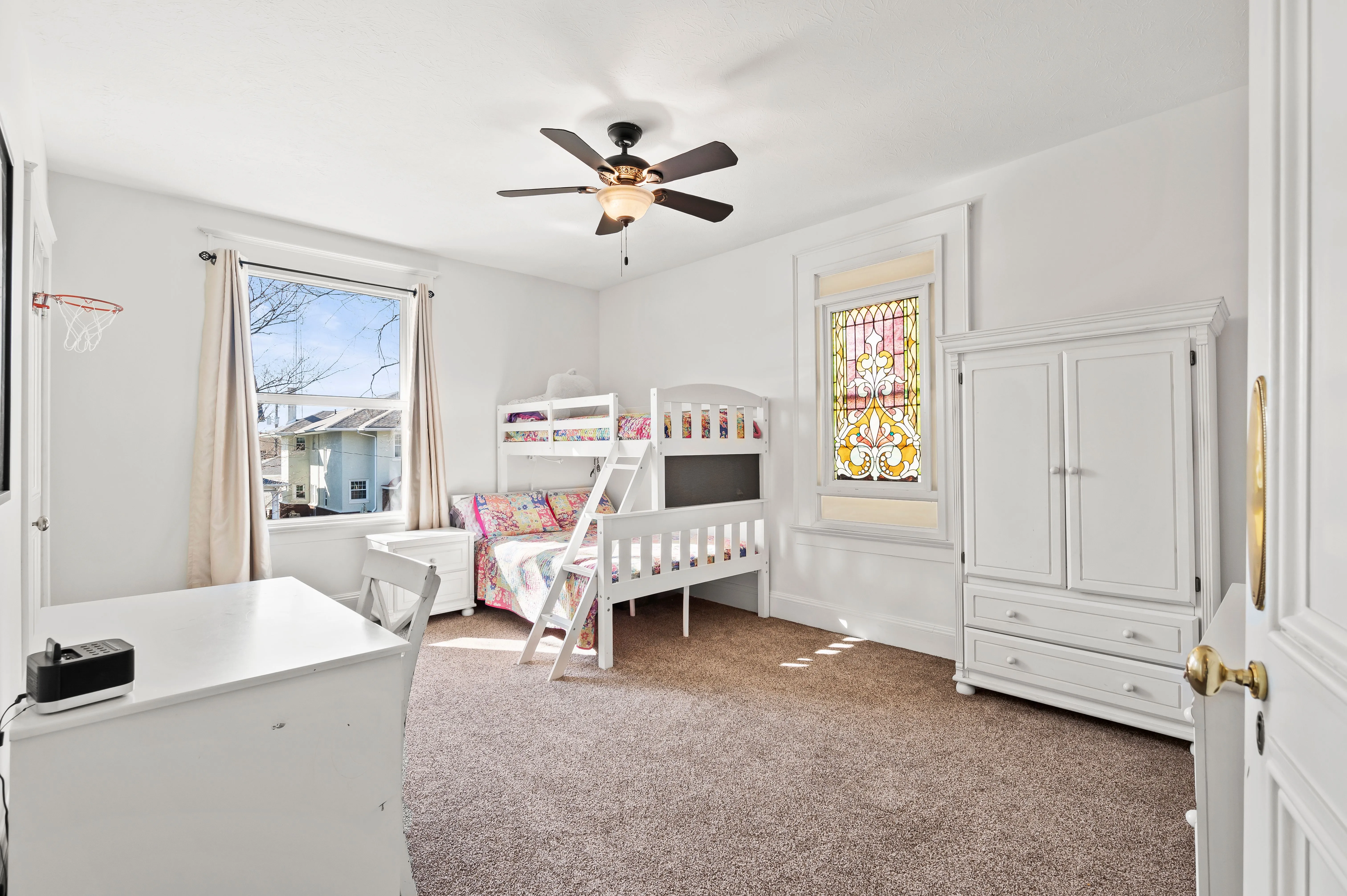 Bright child's bedroom with white bunk beds, a play area, a ceiling fan, and a stained glass window.