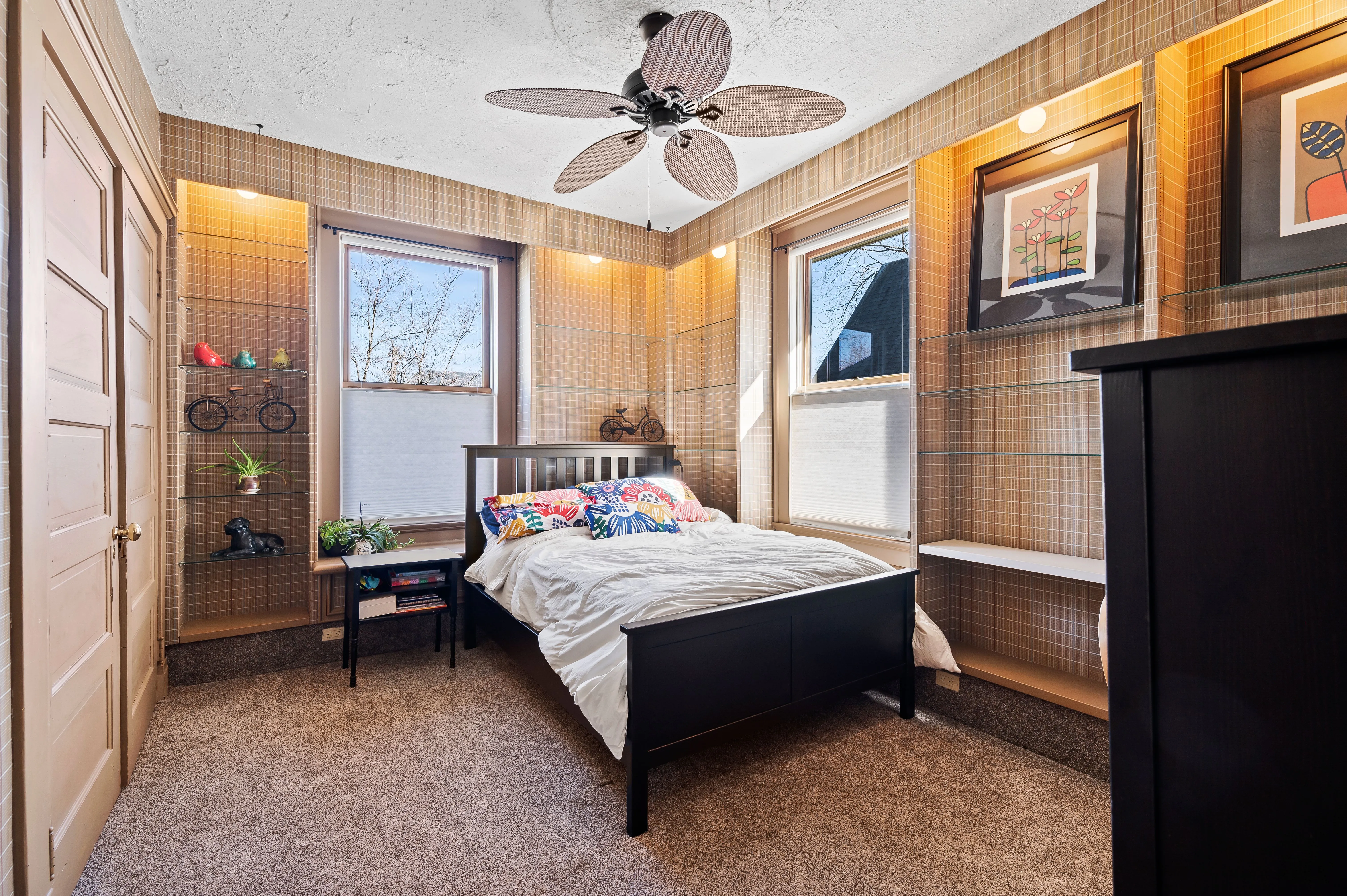 Cozy bedroom with a double bed, side table, ceiling fan, and framed pictures on wood-paneled walls, featuring a carpeted floor and multiple windows.