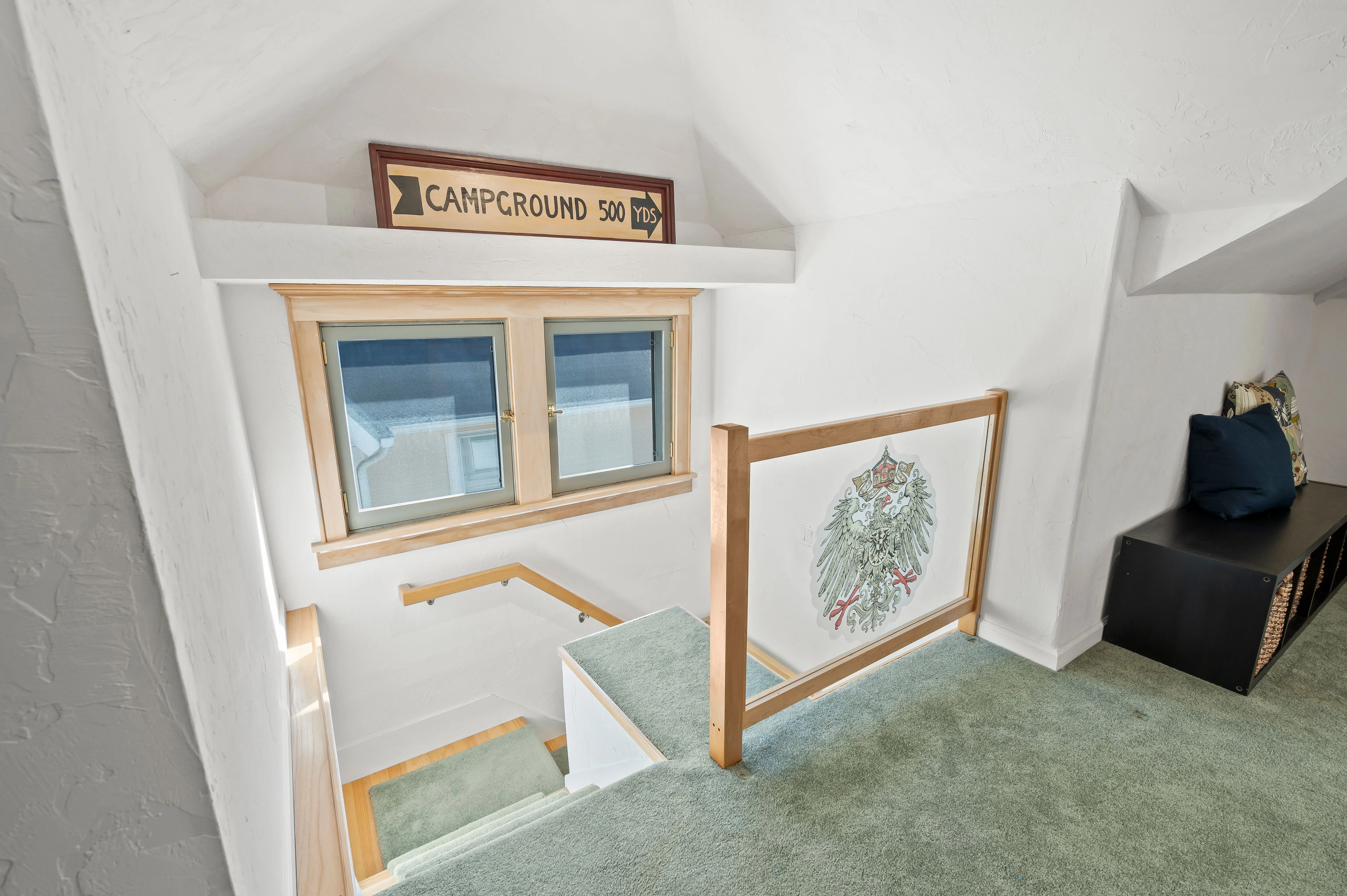Interior view of a cozy attic room with a sign saying "CAMPING ⛺️", a protective railing, two windows, a framed map on the wall, and a black piece of luggage.