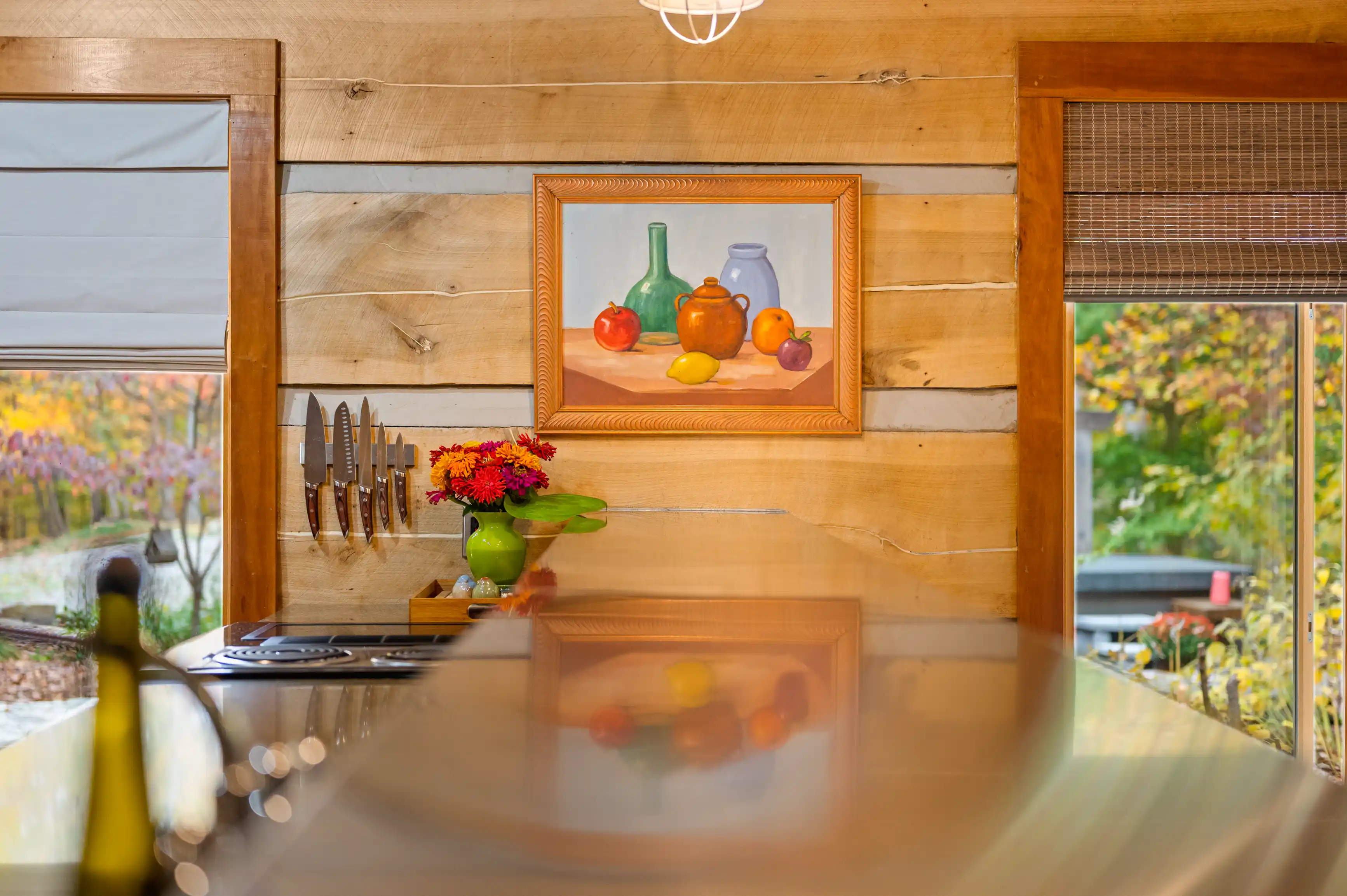 Warm-toned rustic kitchen with a framed still-life painting, knife set on wood wall, red flowers in a green vase, and autumn foliage visible through the window.