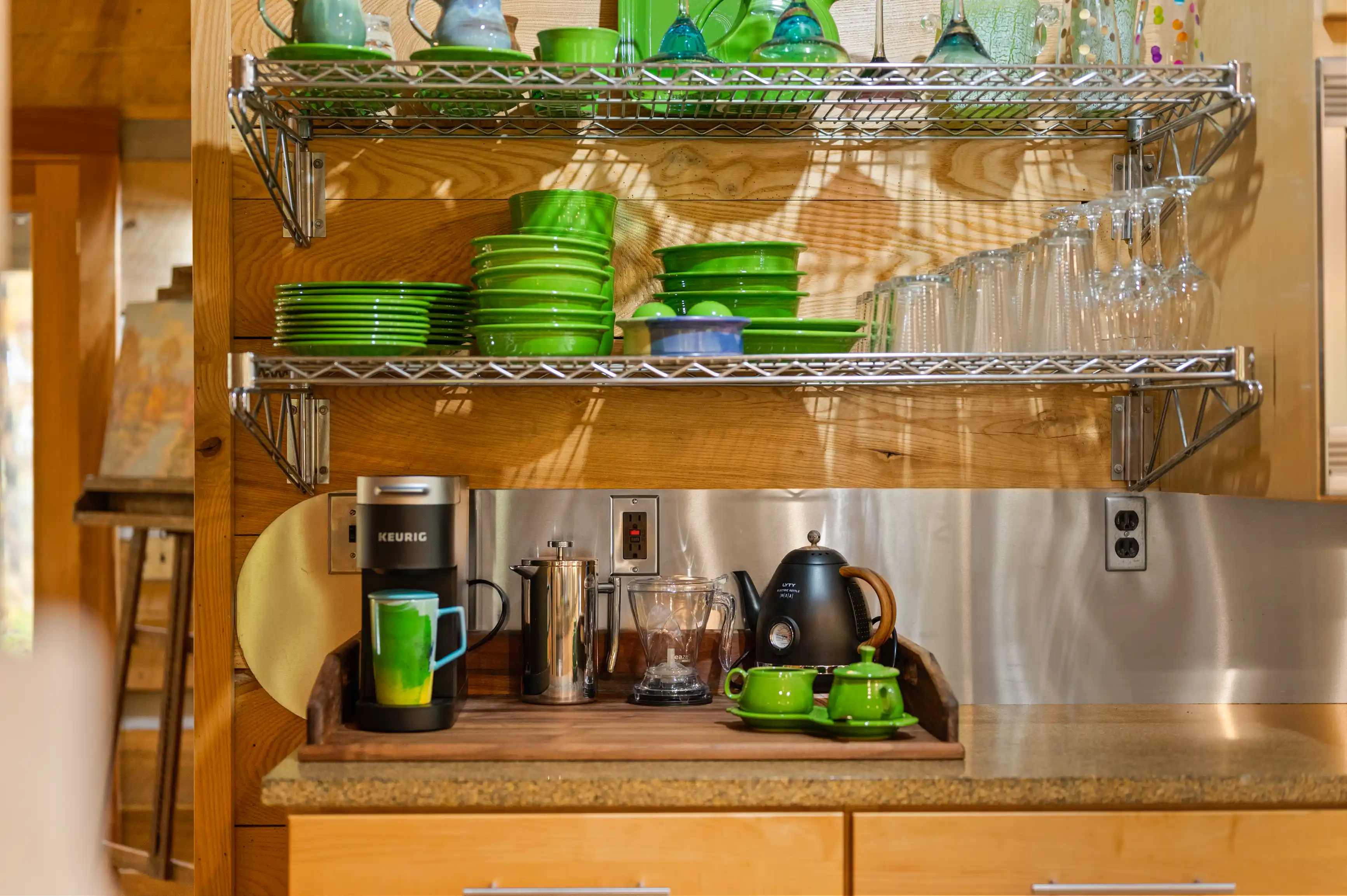 Alt text: A cozy kitchen corner with a wooden counter holding a Keurig coffee maker, French press, and tea pot, with shelves above displaying green plates and glasses.