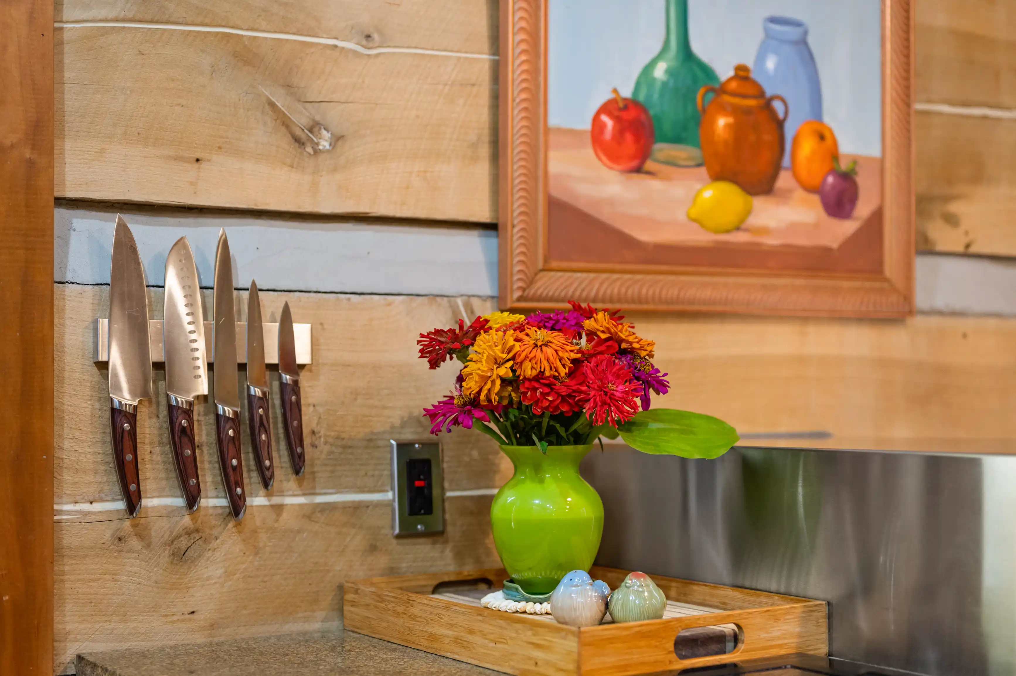 Rustic kitchen setting with a colorful flower bouquet in a green vase, a set of knives on a magnetic strip, and a framed painting of pottery and fruit.