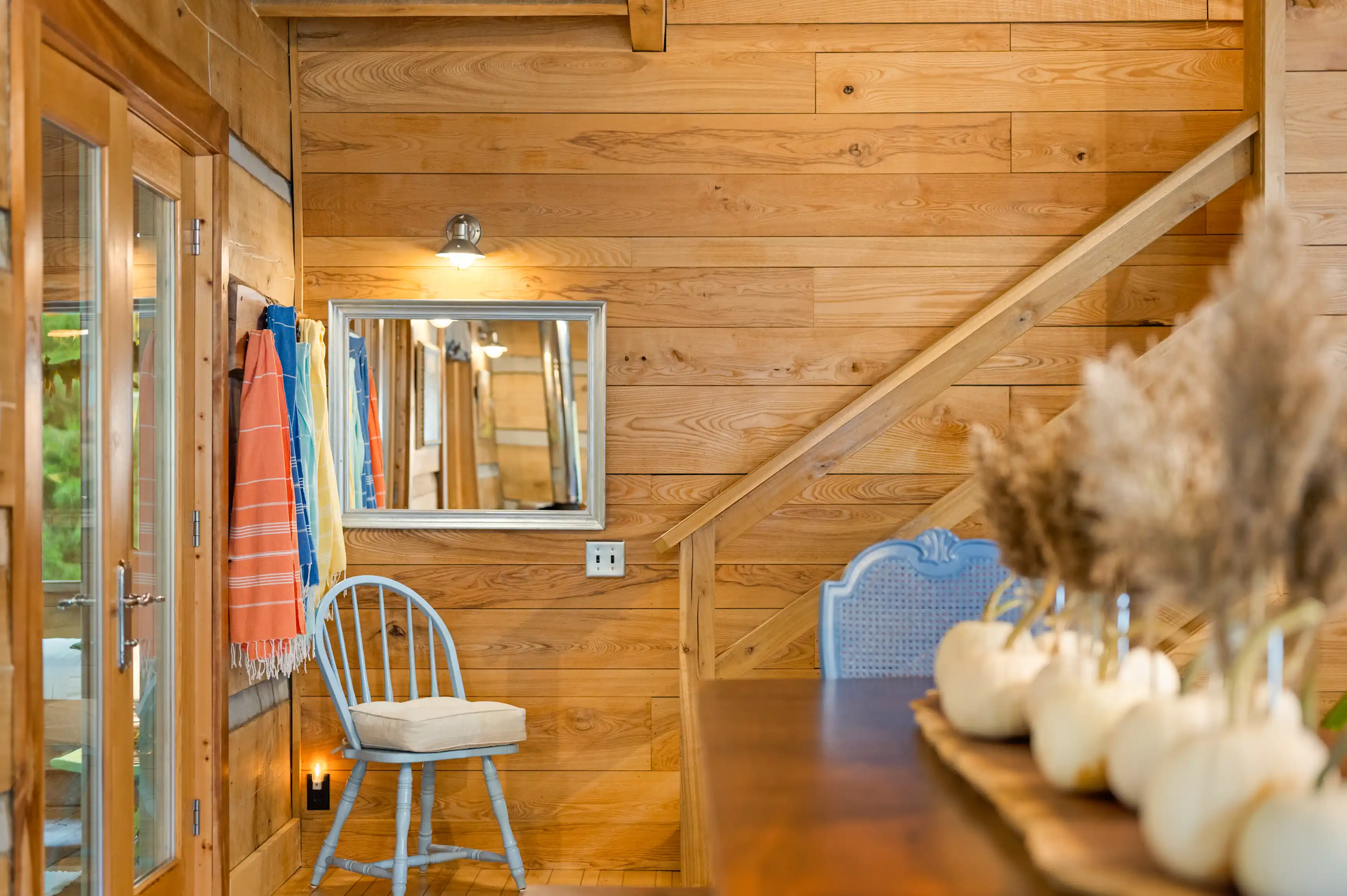 Cozy wooden cabin interior with a staircase, decorative white pumpkins, dried pampas grass arrangement, and colorful towels hanging by the glass door.
