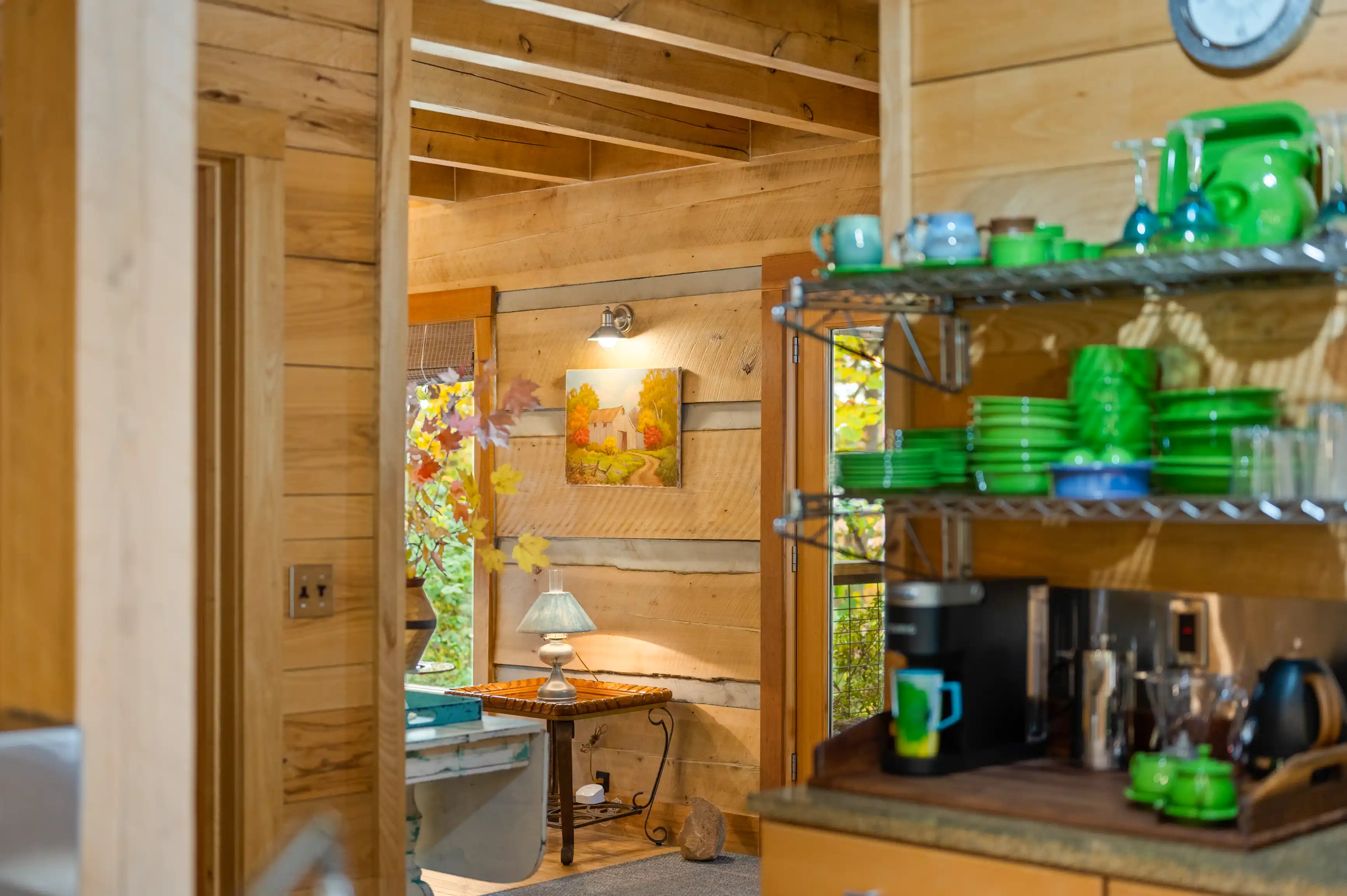 Cozy wooden cabin interior with a focus on a well-organized kitchen shelf stocked with green ceramics and silver appliances, adjacent to a warm sitting area with a lamp and a small table, all bathed in natural light filtering through autumn leaves visible through a window.