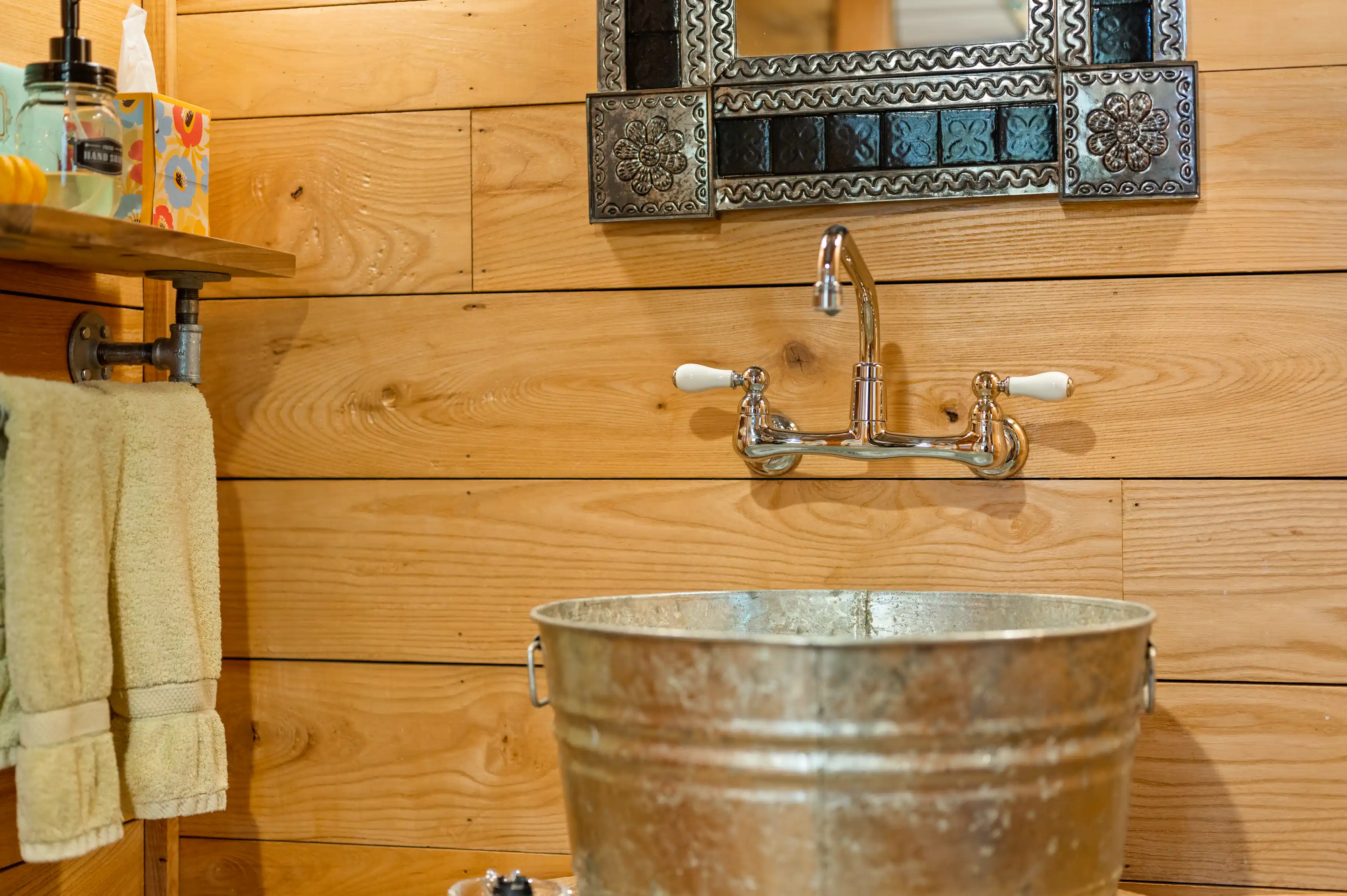 Rustic bathroom interior with a galvanized bucket sink, wall-mounted faucet, wooden walls and a mirror with decorative metal frame.