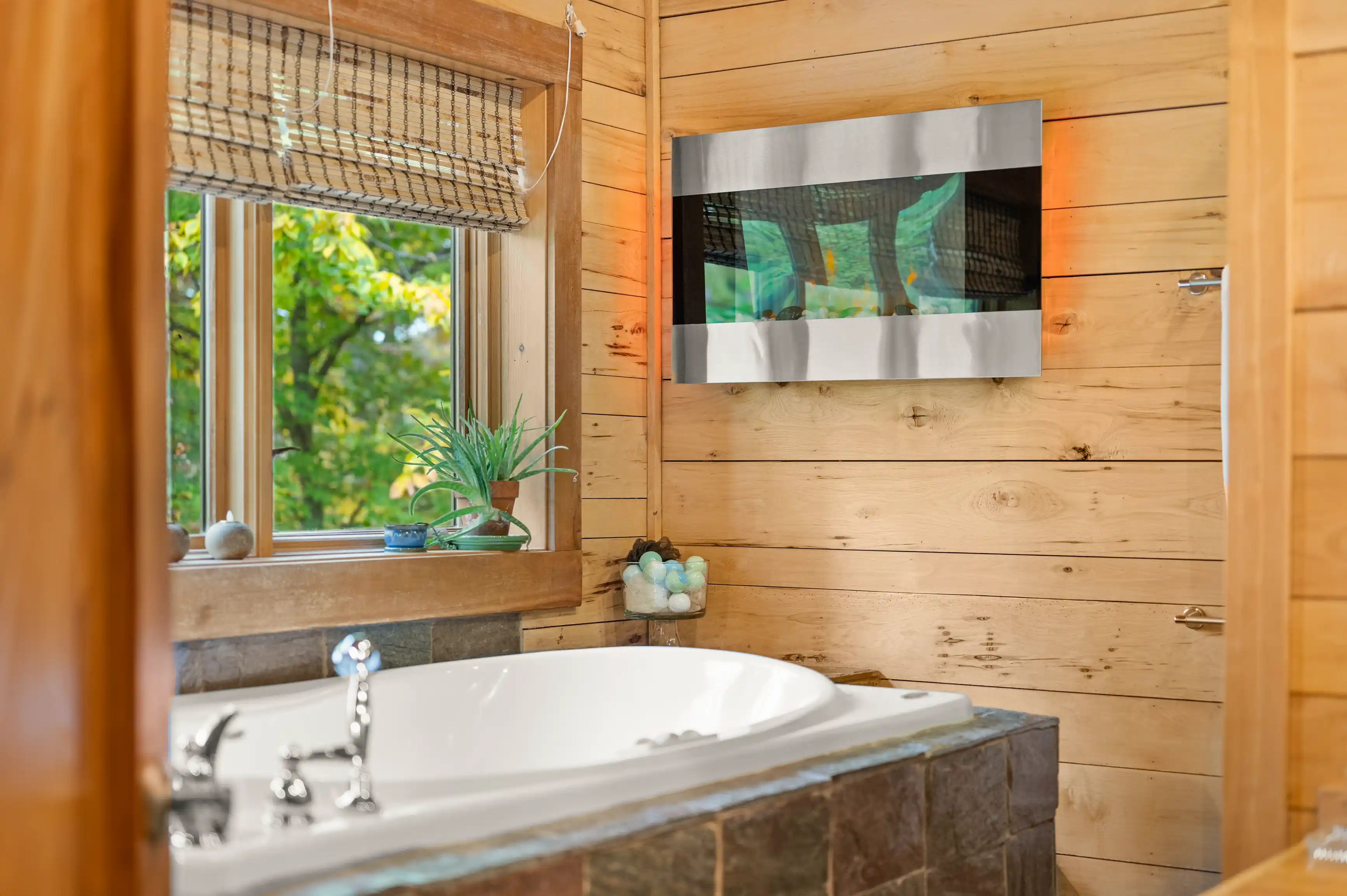 Interior of a rustic bathroom with a wooden bathtub surround, matching wooden walls, a plant on the windowsill, and a modern mirror above the tub.