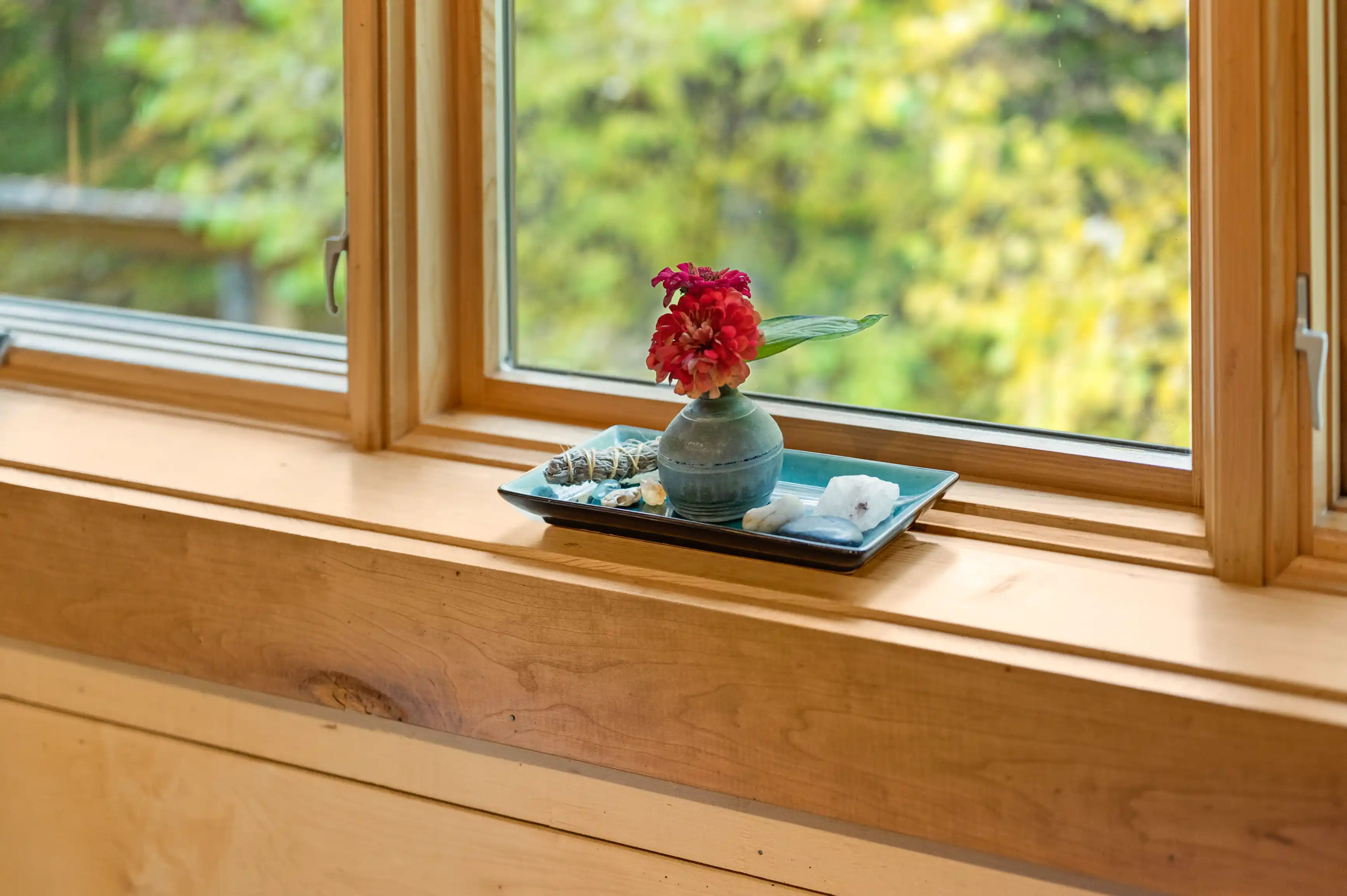 A small vase with a red flower and some stones on a blue tray by a wooden window, with a lush greenery view outside.