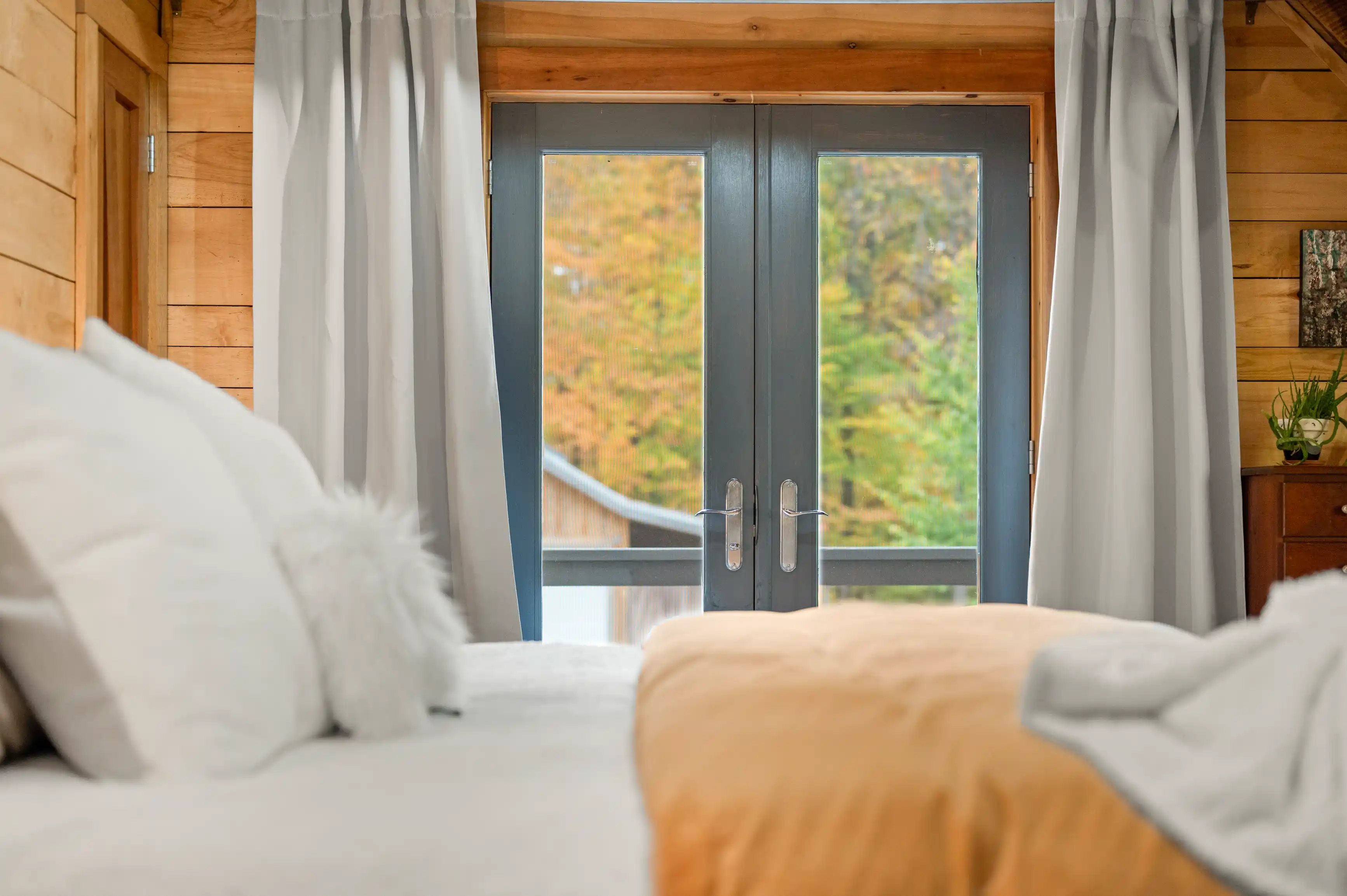 A cozy bedroom with a view of autumn foliage through a glass door, flanked by grey curtains.