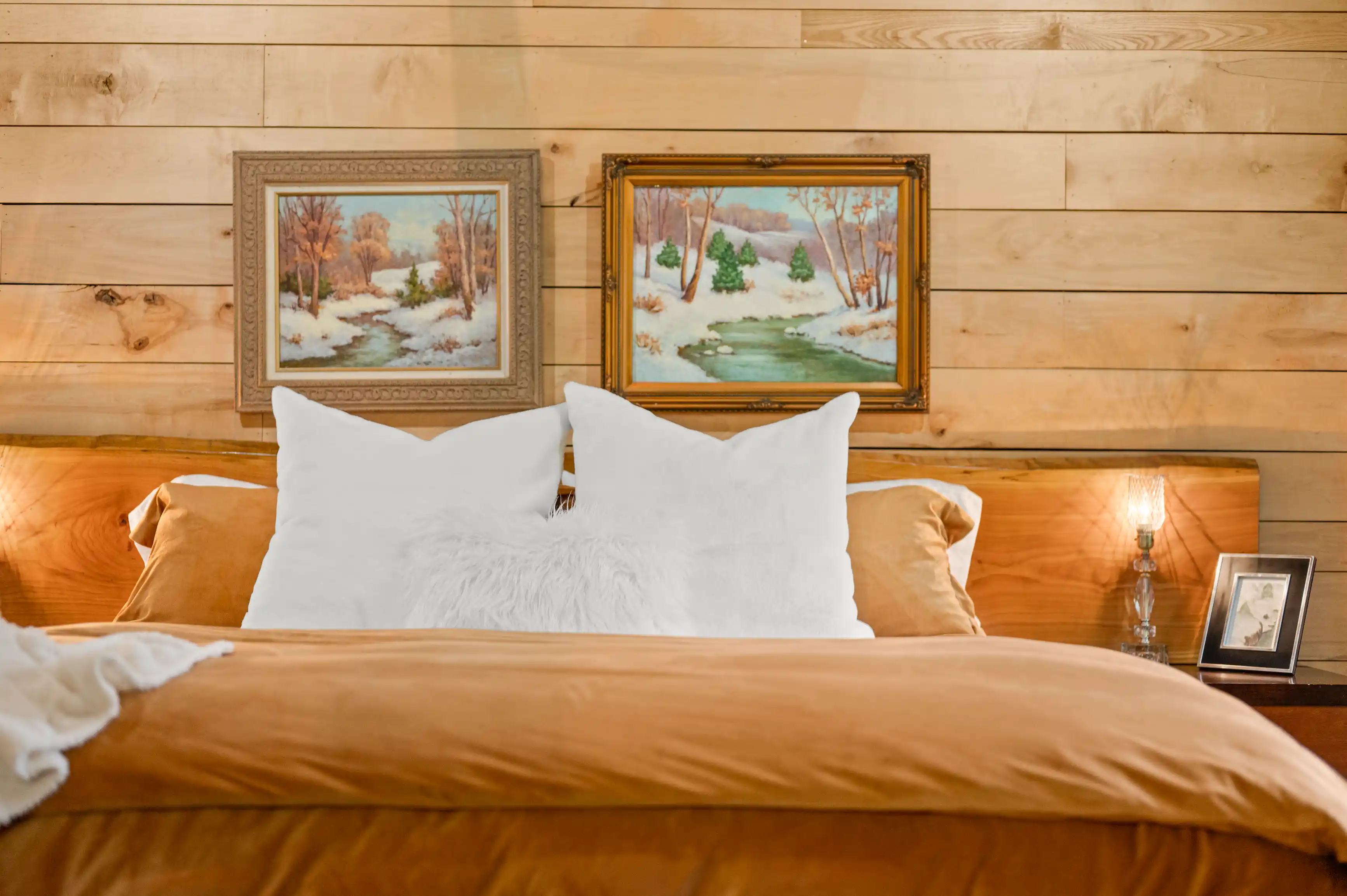 A cozy bedroom with a wooden headboard, warm-toned bedding, white fluffy pillows, and two framed landscape paintings above the bed.