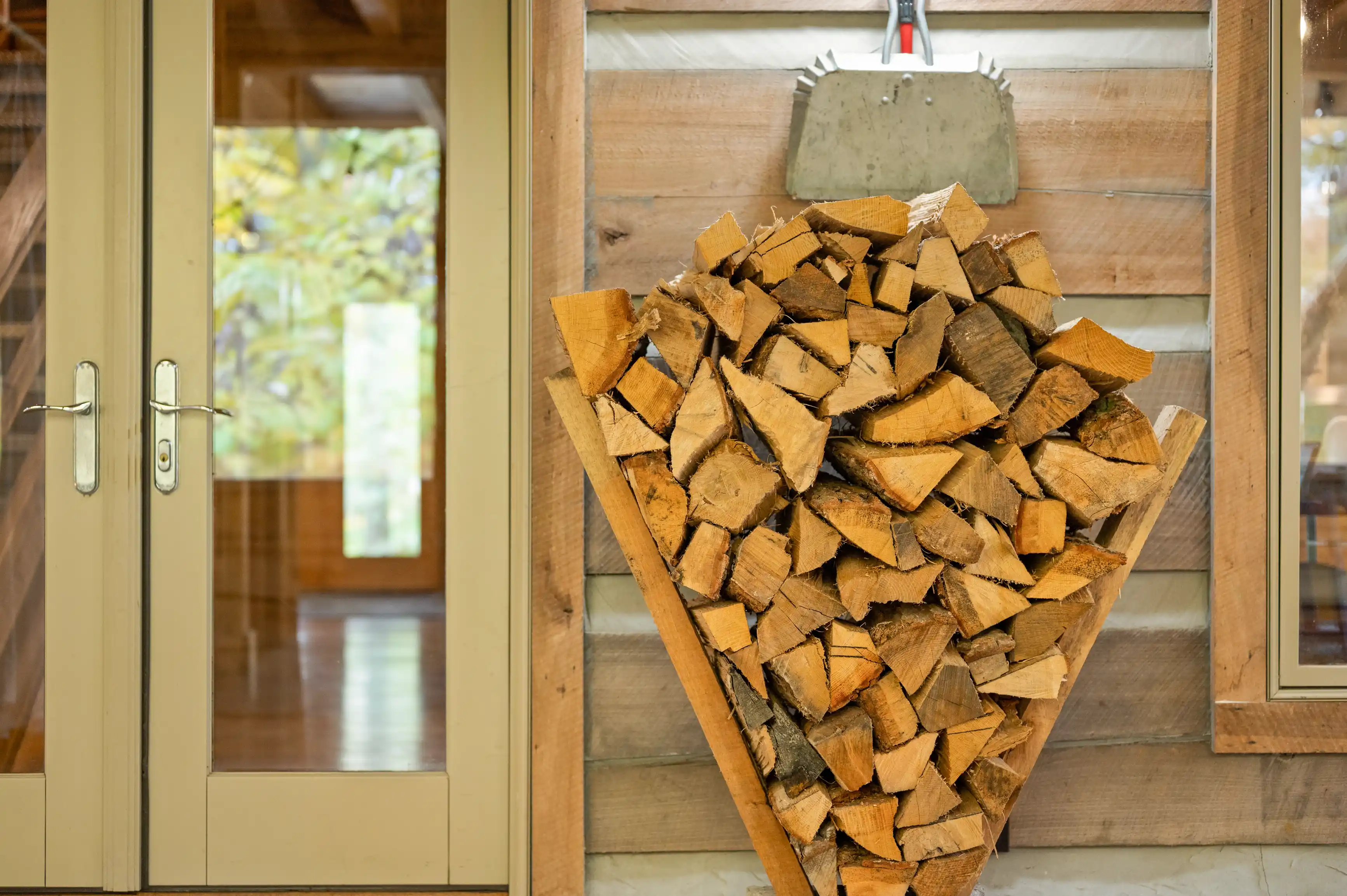 A neatly stacked pile of firewood contained within a wooden holder against a wall next to glass doors.