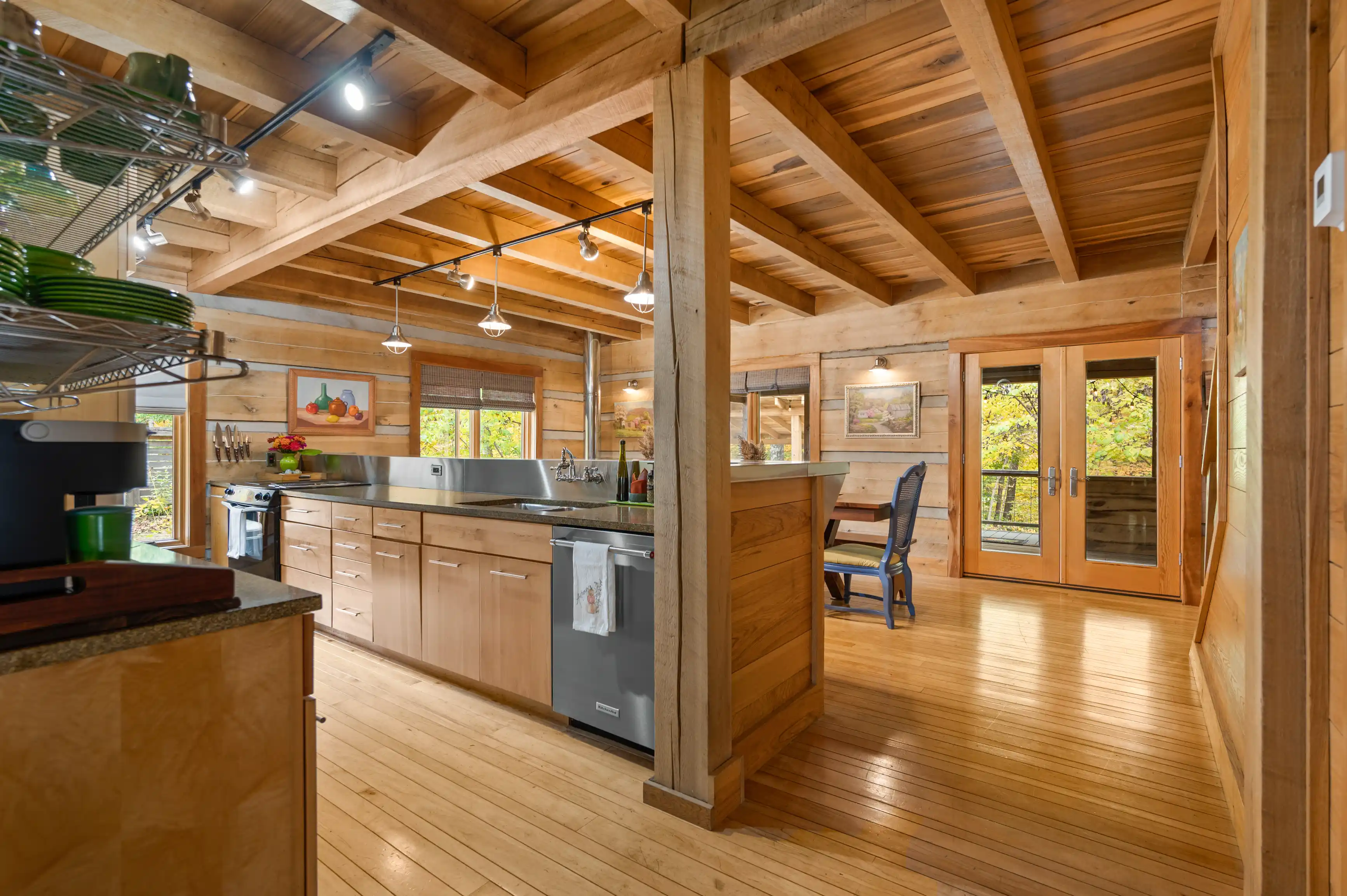 Rustic wooden kitchen interior with exposed beams, stainless steel appliances, and pendant lighting with a view of trees through glass doors.