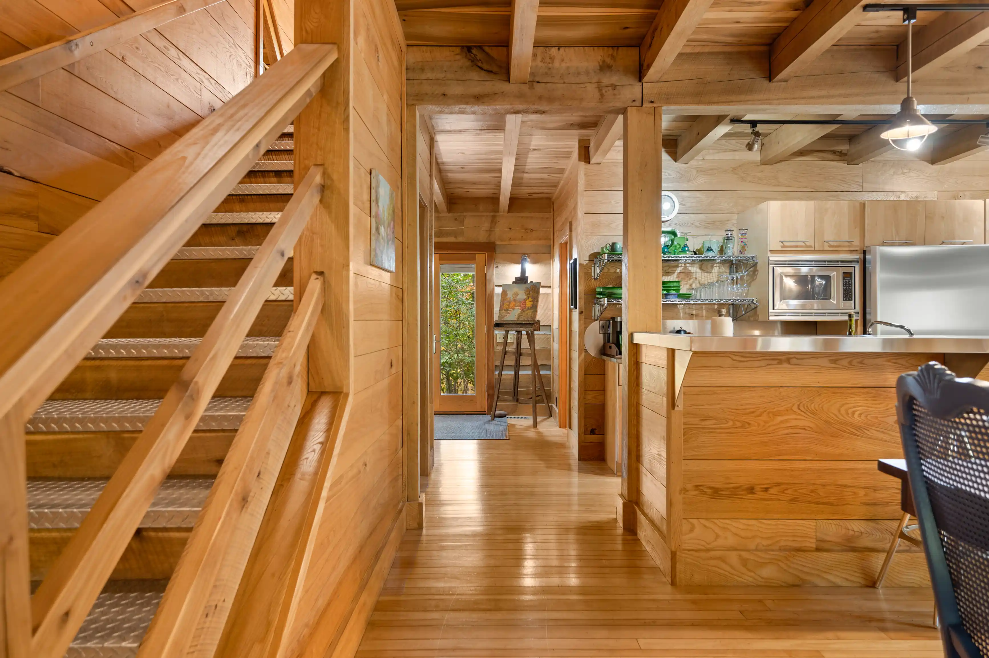 Interior of a cozy wooden cabin featuring a staircase on the left, a kitchen with modern appliances to the right, and a glimpse of a bright doorway ahead.
