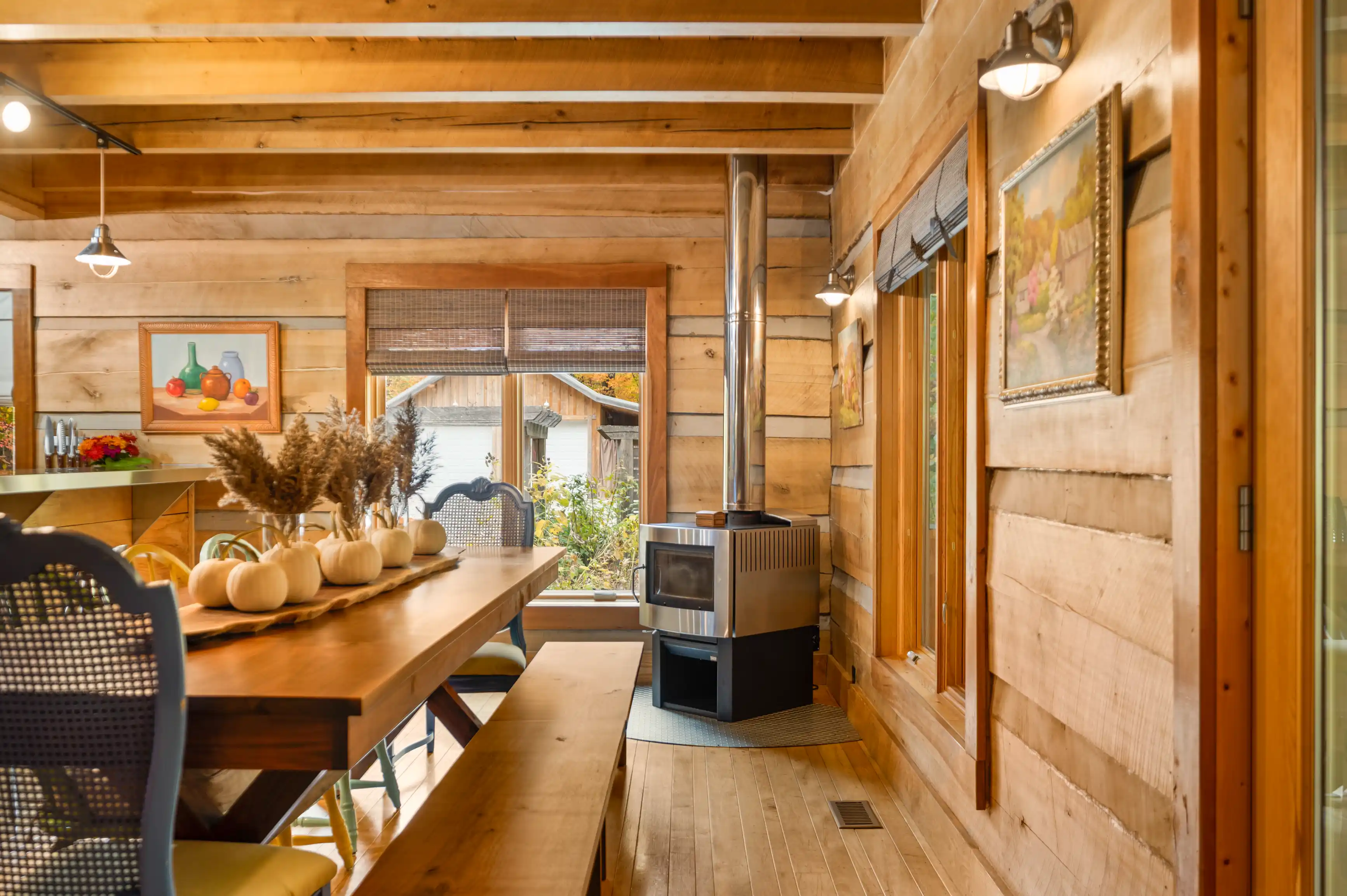 Cozy wooden cabin interior with a long dining table adorned with pumpkins, a wood stove, framed art on walls, and a view of the outdoors through windows.