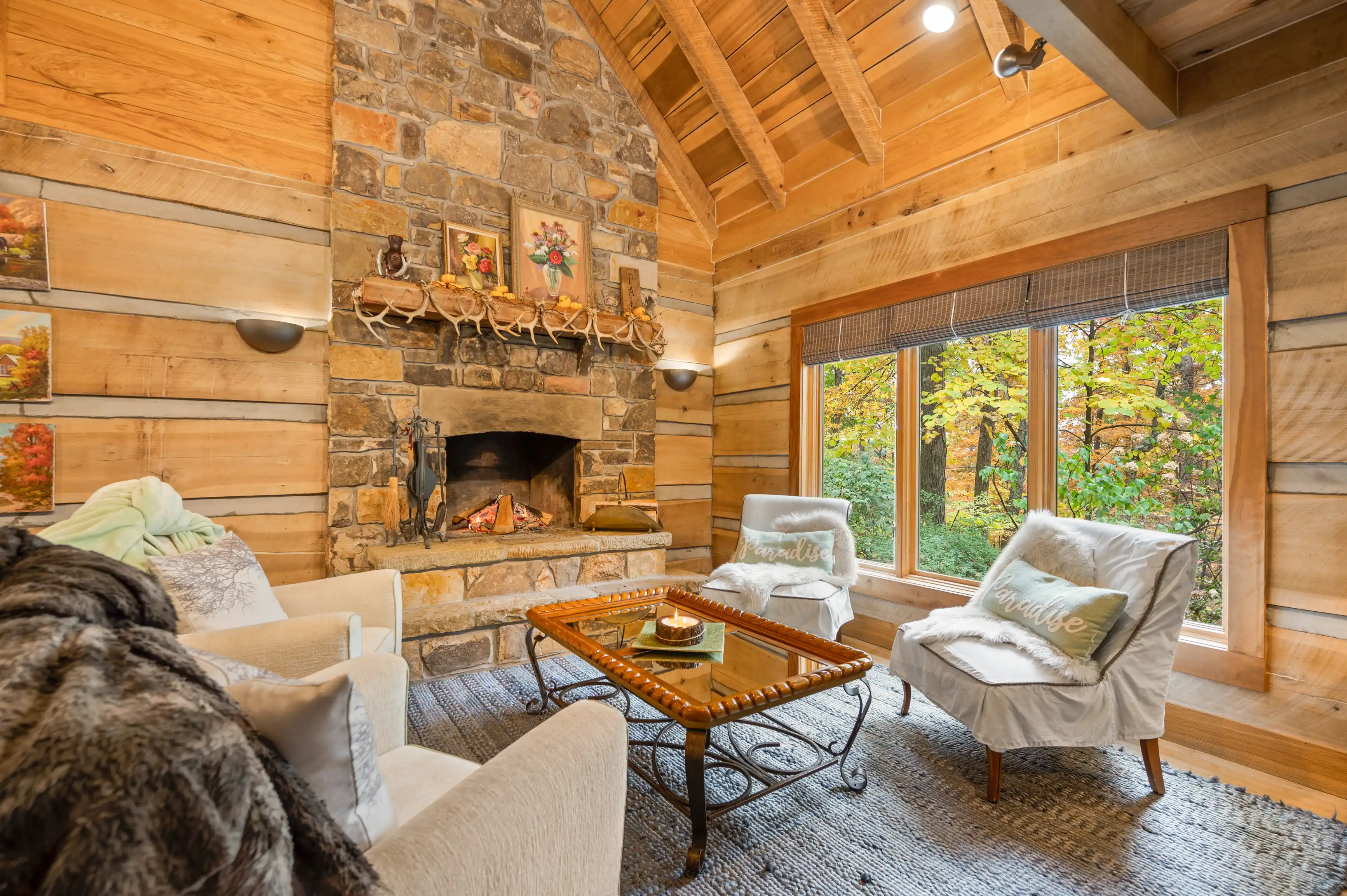 Cozy cabin living room interior with a stone fireplace, wooden walls and ceiling, rustic furniture, and large windows with a forest view.