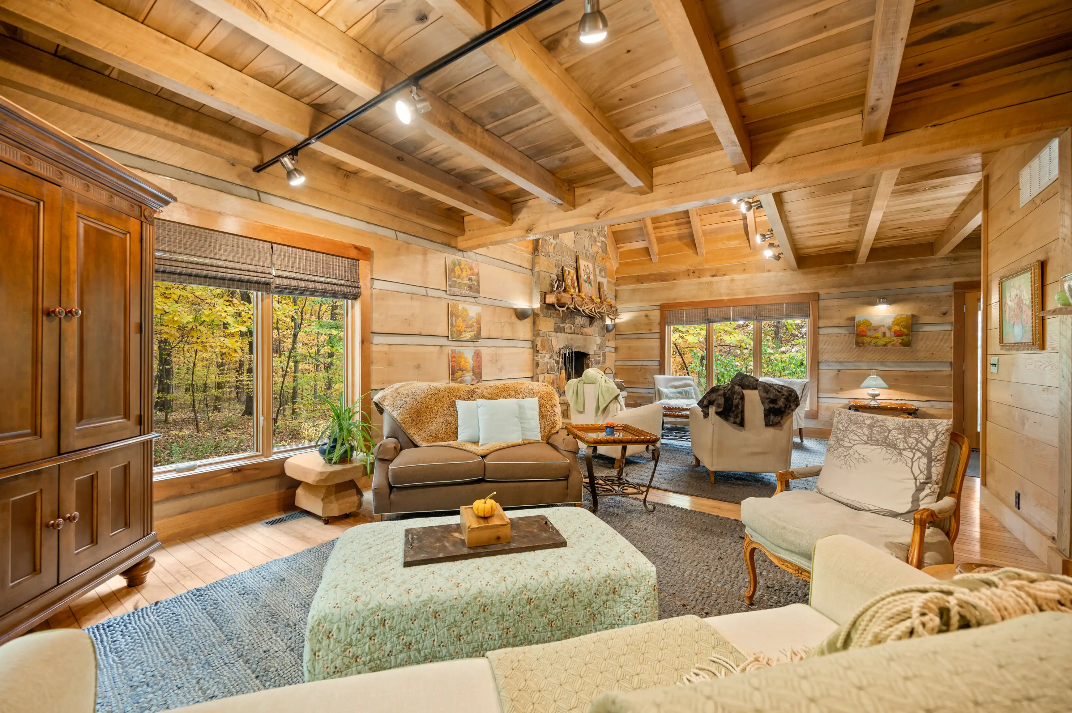 Cozy rustic living room with wooden walls, exposed beams, a stone fireplace, and large windows with a forest view.