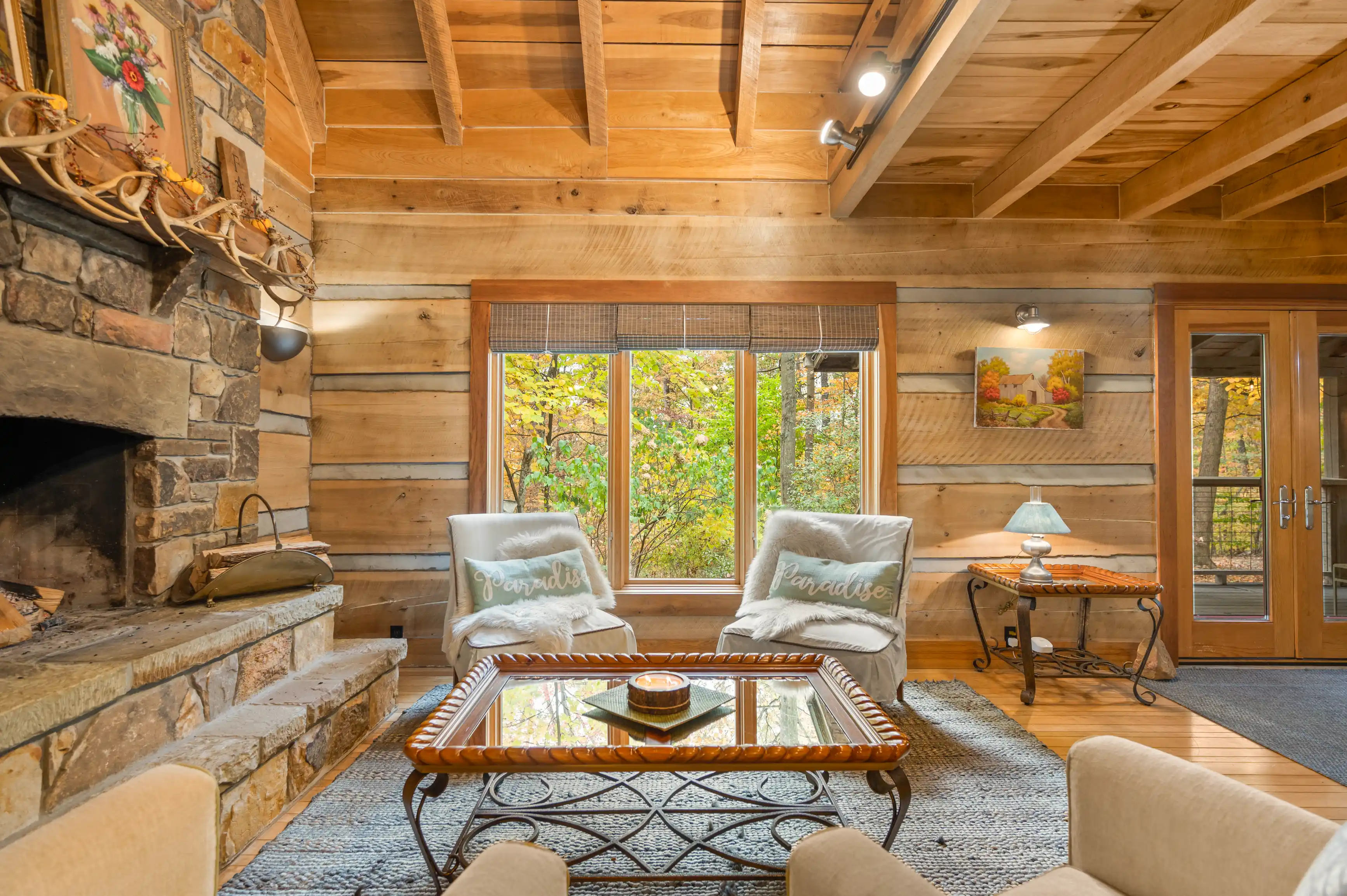 Cozy cabin interior with a stone fireplace, wooden walls, exposed beams, and a view of the autumn trees through a large window.