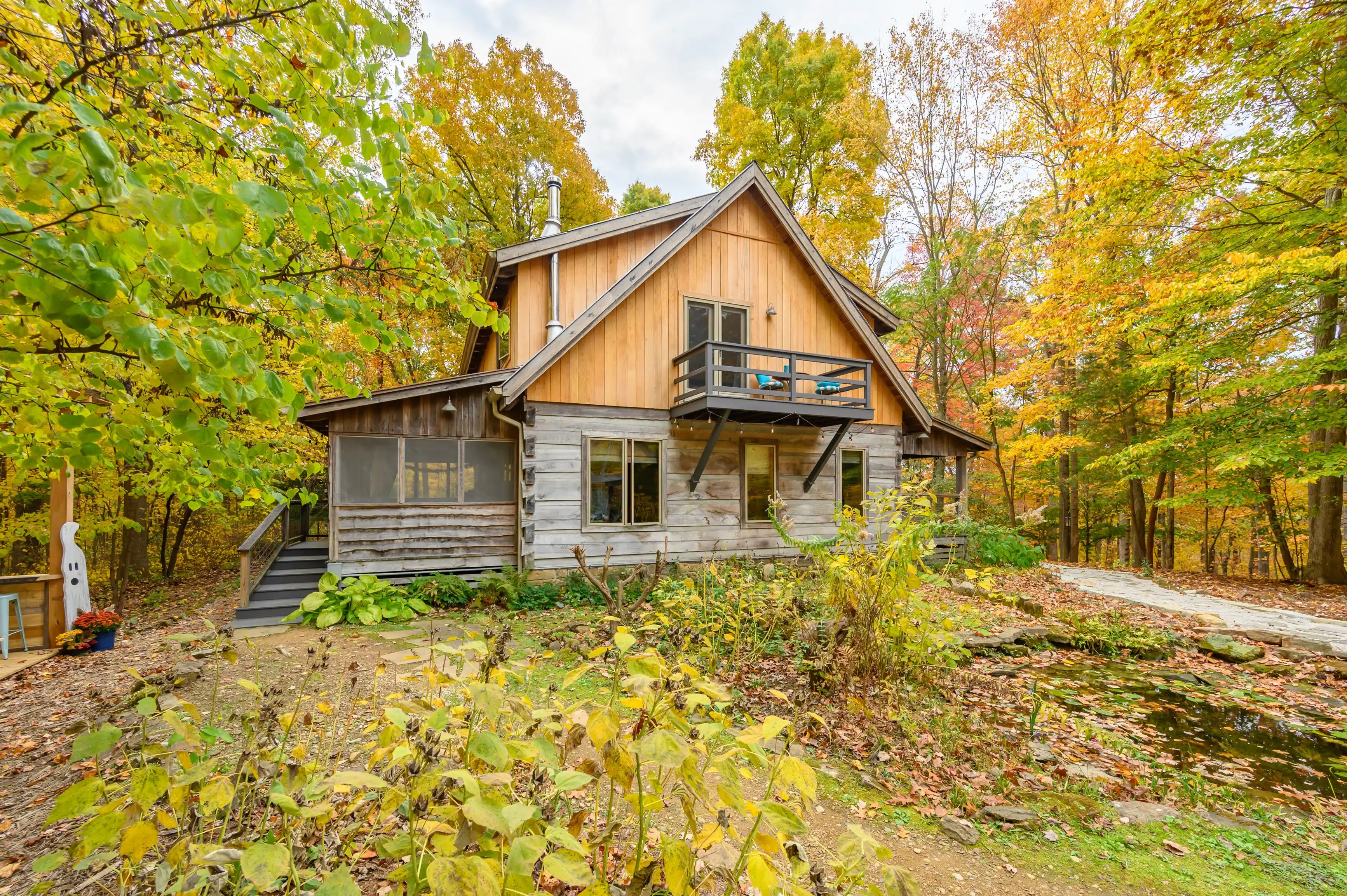 A cozy wooden cabin surrounded by autumn-colored trees with a balcony and a pond in the forefront.