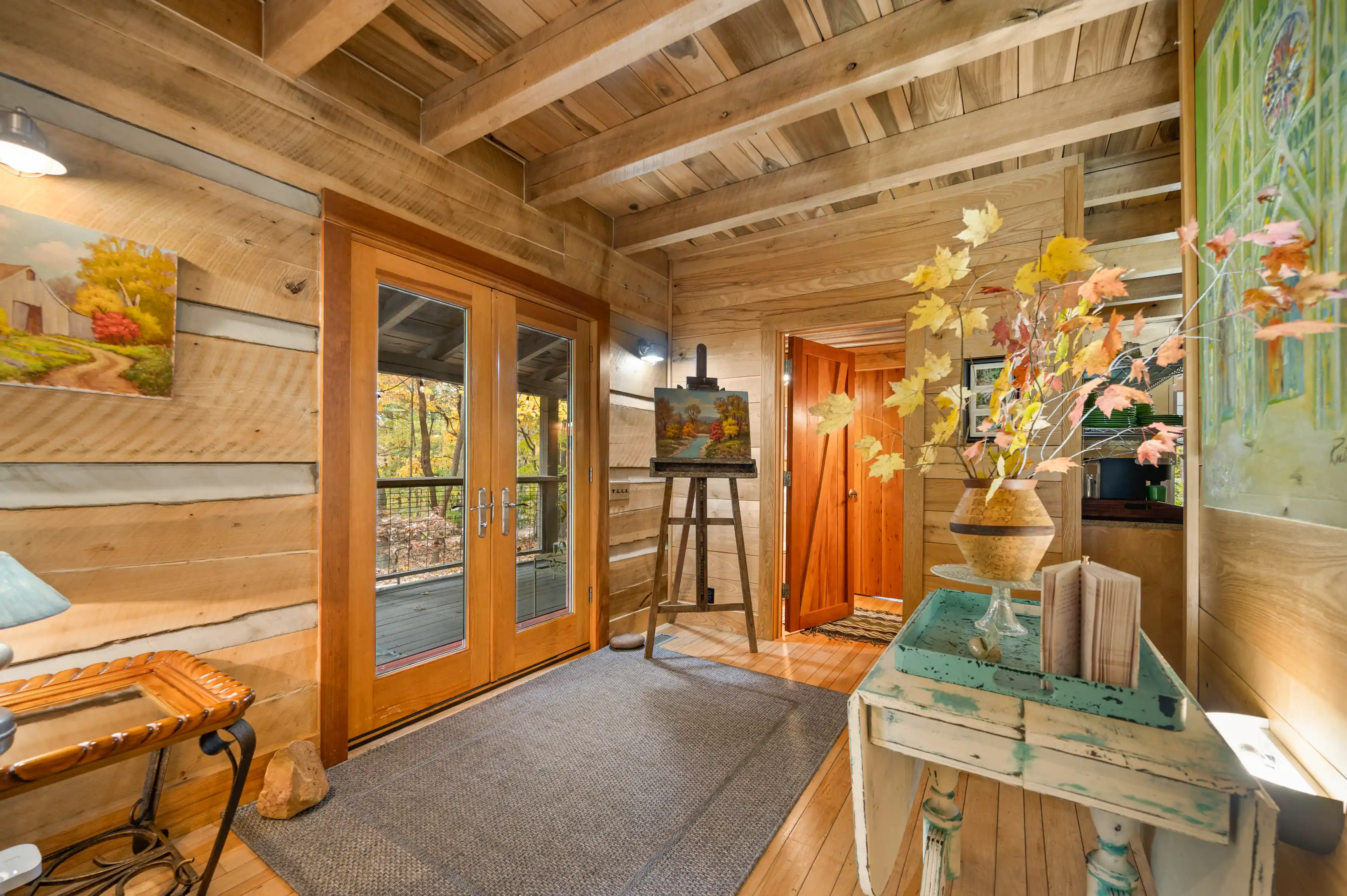 Interior of a cozy wooden cabin with autumn-inspired decorations, featuring a large glass door, a painting on an easel, and a rustic table with a vase of fall leaves.