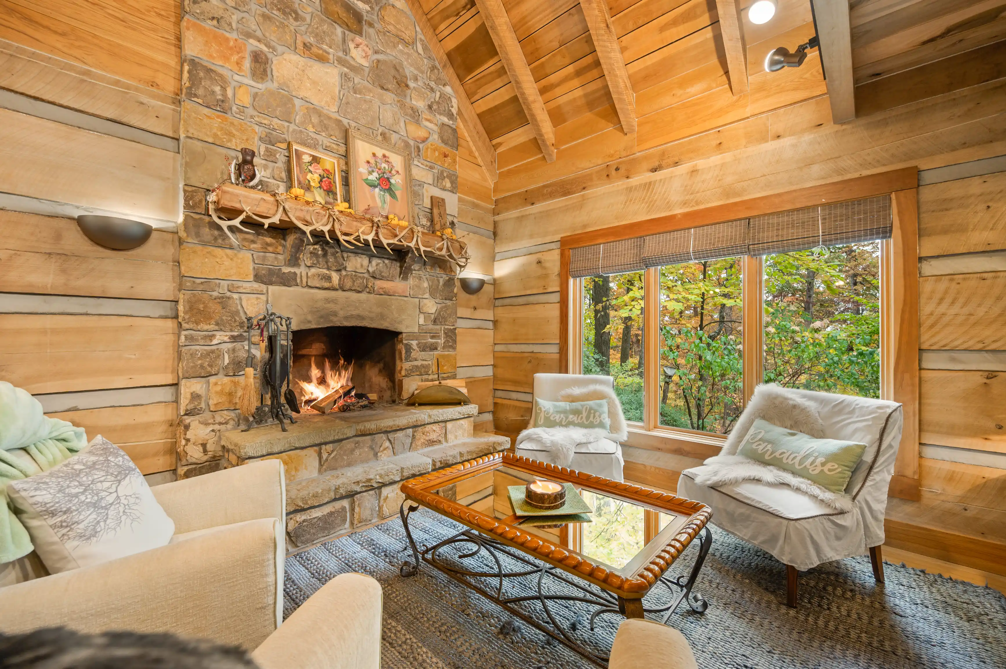 Cozy cabin living room with a stone fireplace, wooden walls, beamed ceiling, and a window view of autumn foliage.