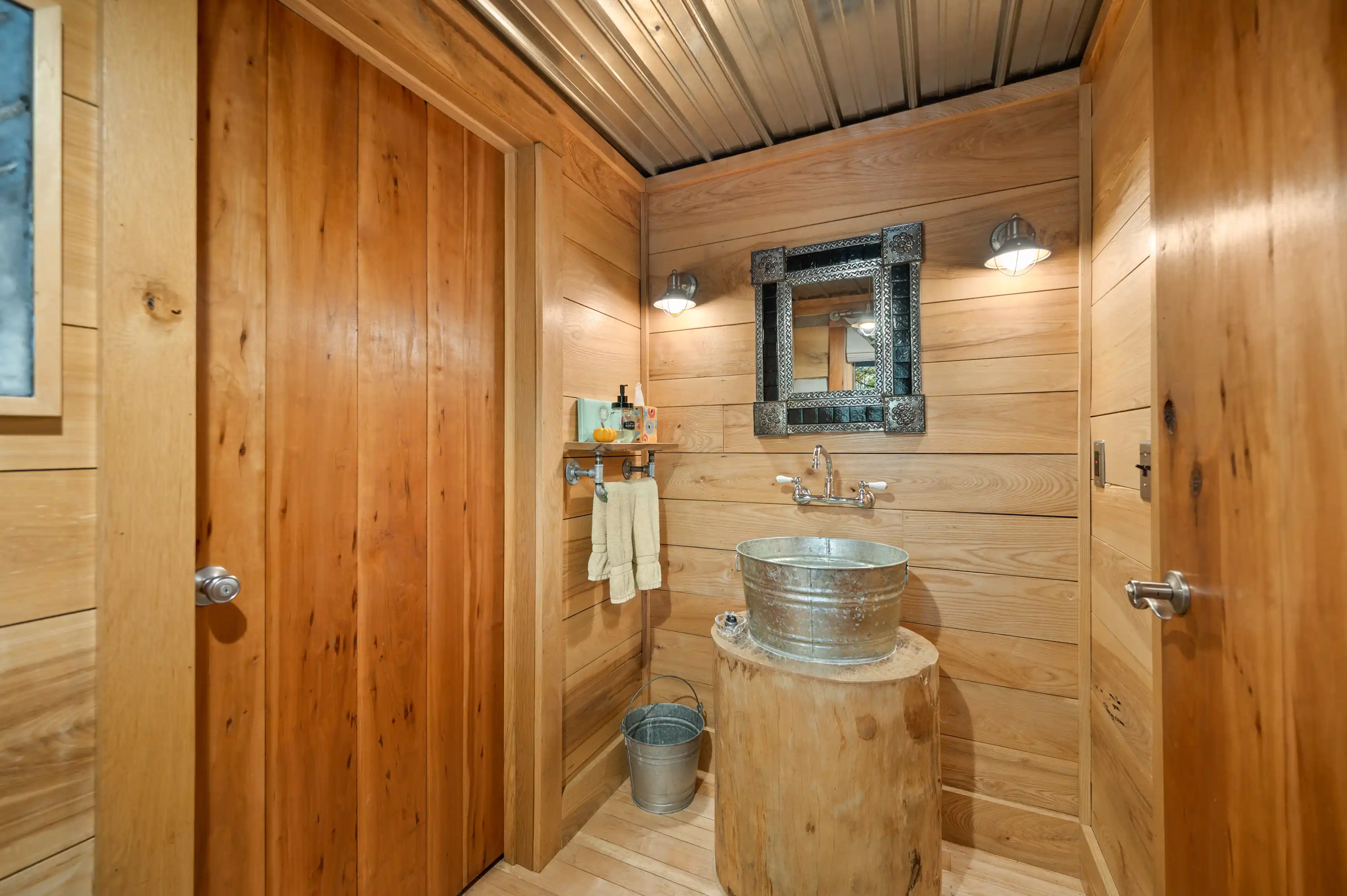 Rustic wooden bathroom interior featuring a galvanized bucket sink on a natural wood stump, wall-mounted faucet, framed mirror, and wood-paneled walls.