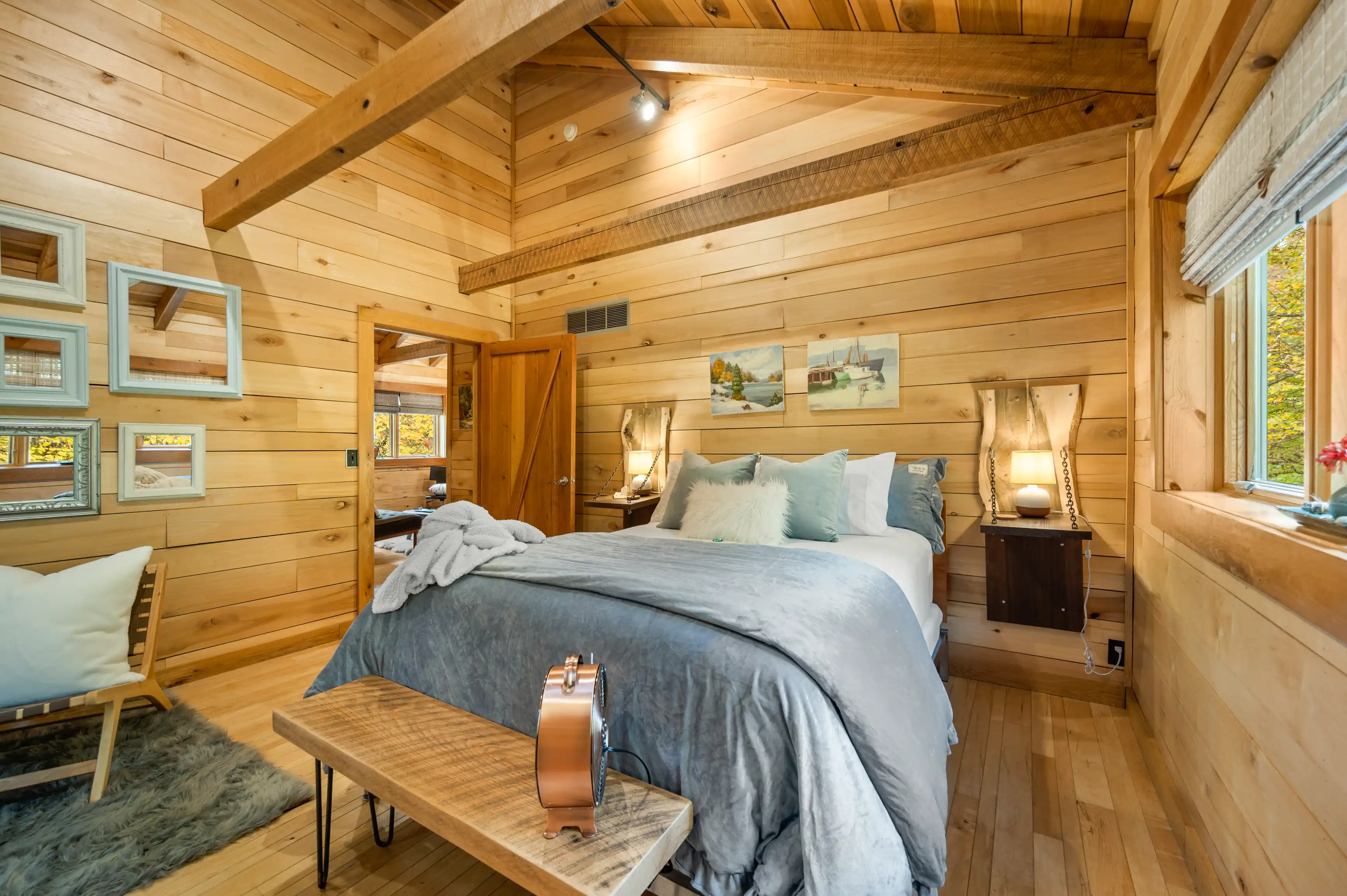 Cozy wooden cabin bedroom interior with a made-up bed, fluffy pillows, hanging artwork, and a warm blanket draped over the corner, with natural light streaming in through the windows.