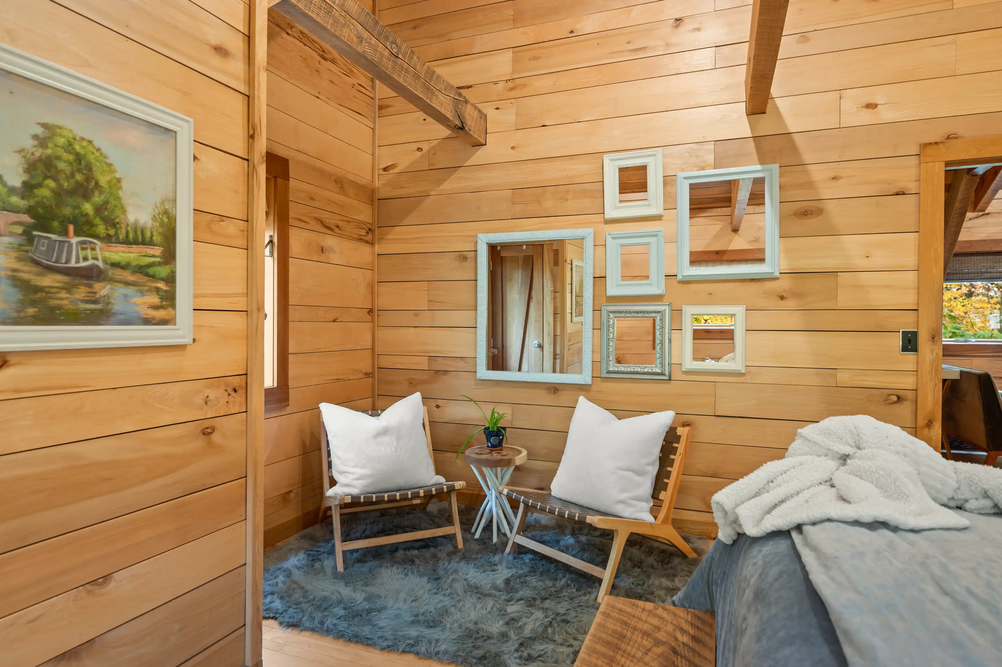 Cozy wooden cabin interior with two chairs, a plush rug, a rustic bed, and decorative mirrors on the wall.