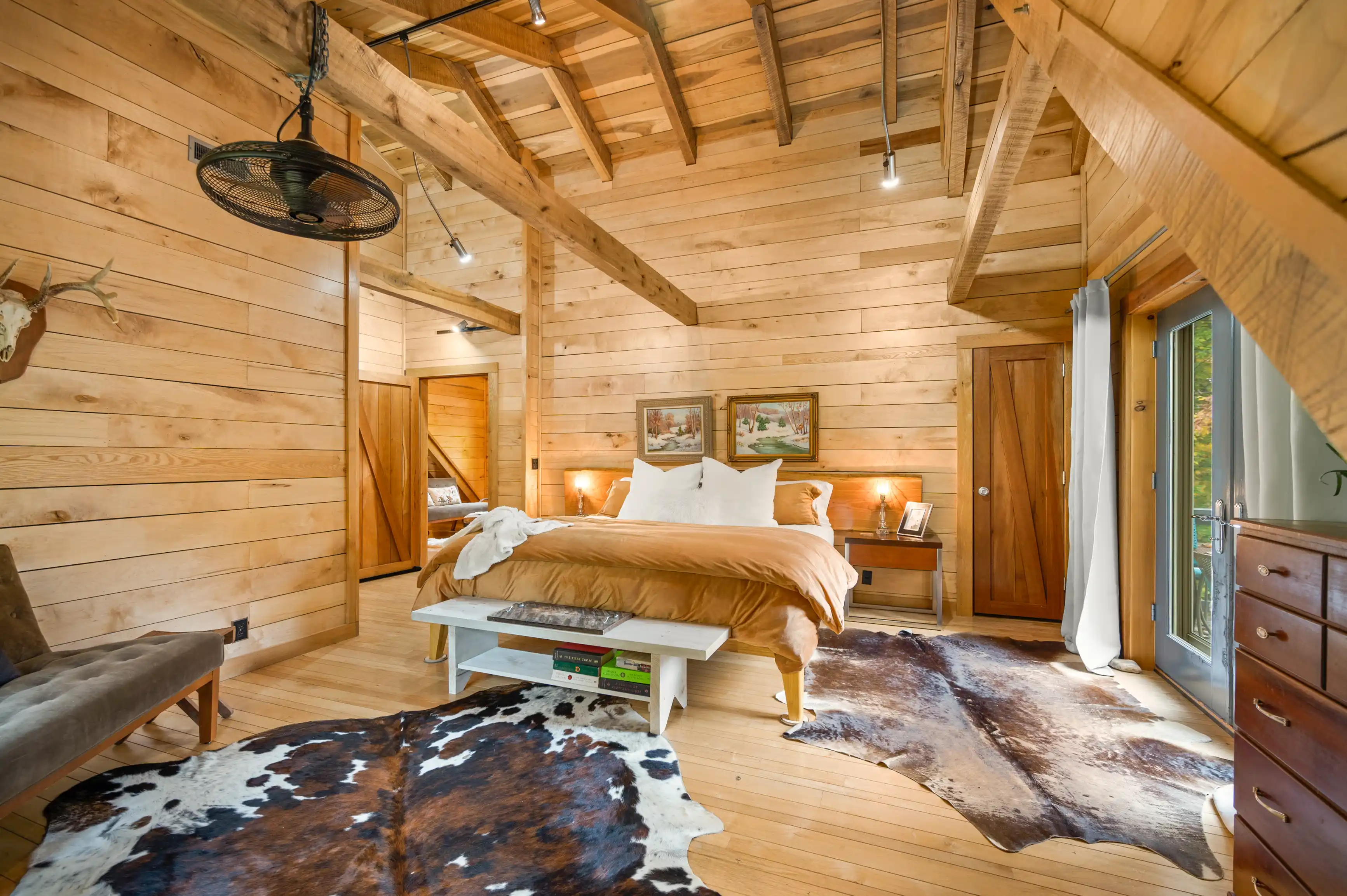 Cozy rustic bedroom with wooden walls and beams, featuring a queen-sized bed with beige bedding, a fur rug on the floor, and a ceiling fan above.