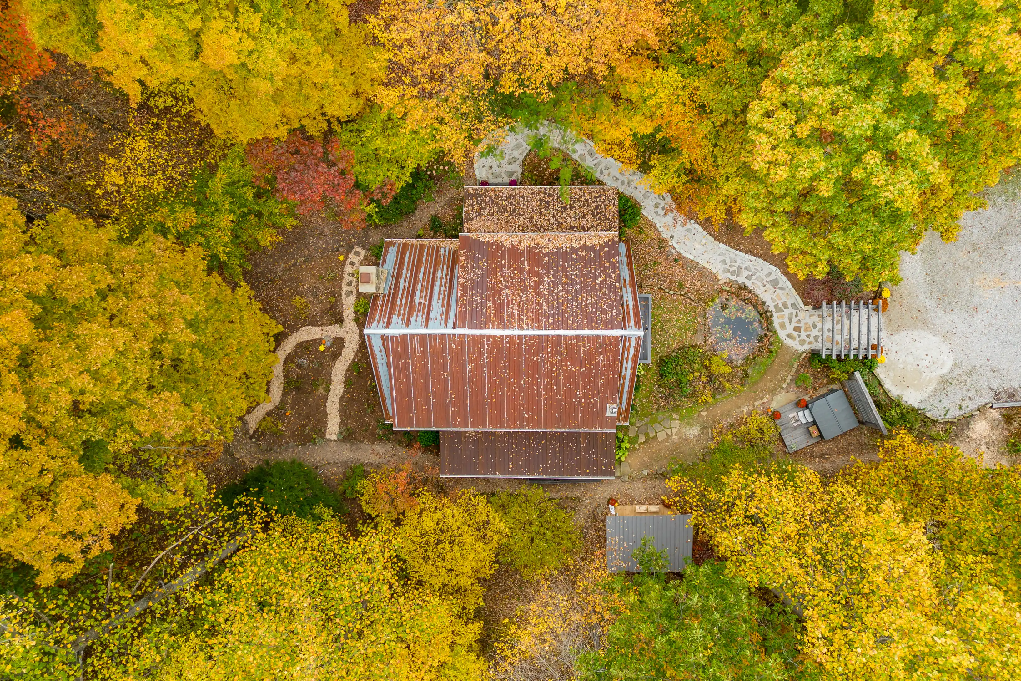 Aerial view of a rustic cabin surrounded by colorful autumn trees with a winding path and a small clearing.